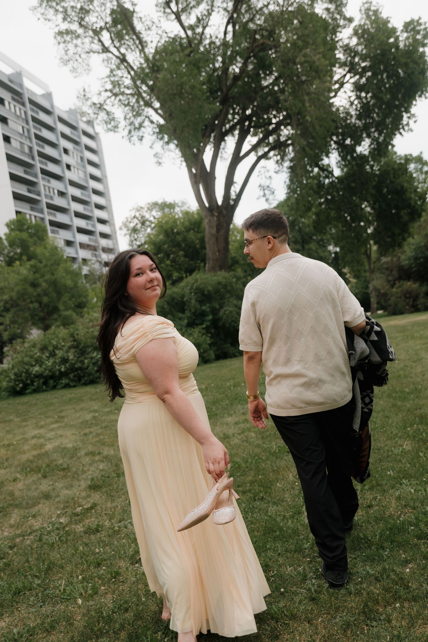 A couple in light-colored outfits pose together on a grassy hillside with apartment buildings visible in the background.