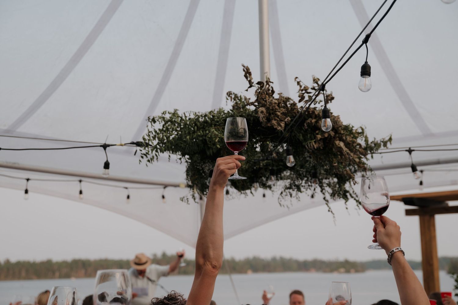 String lights and greenery decorate an outdoor tent for a waterfront wedding celebration.
