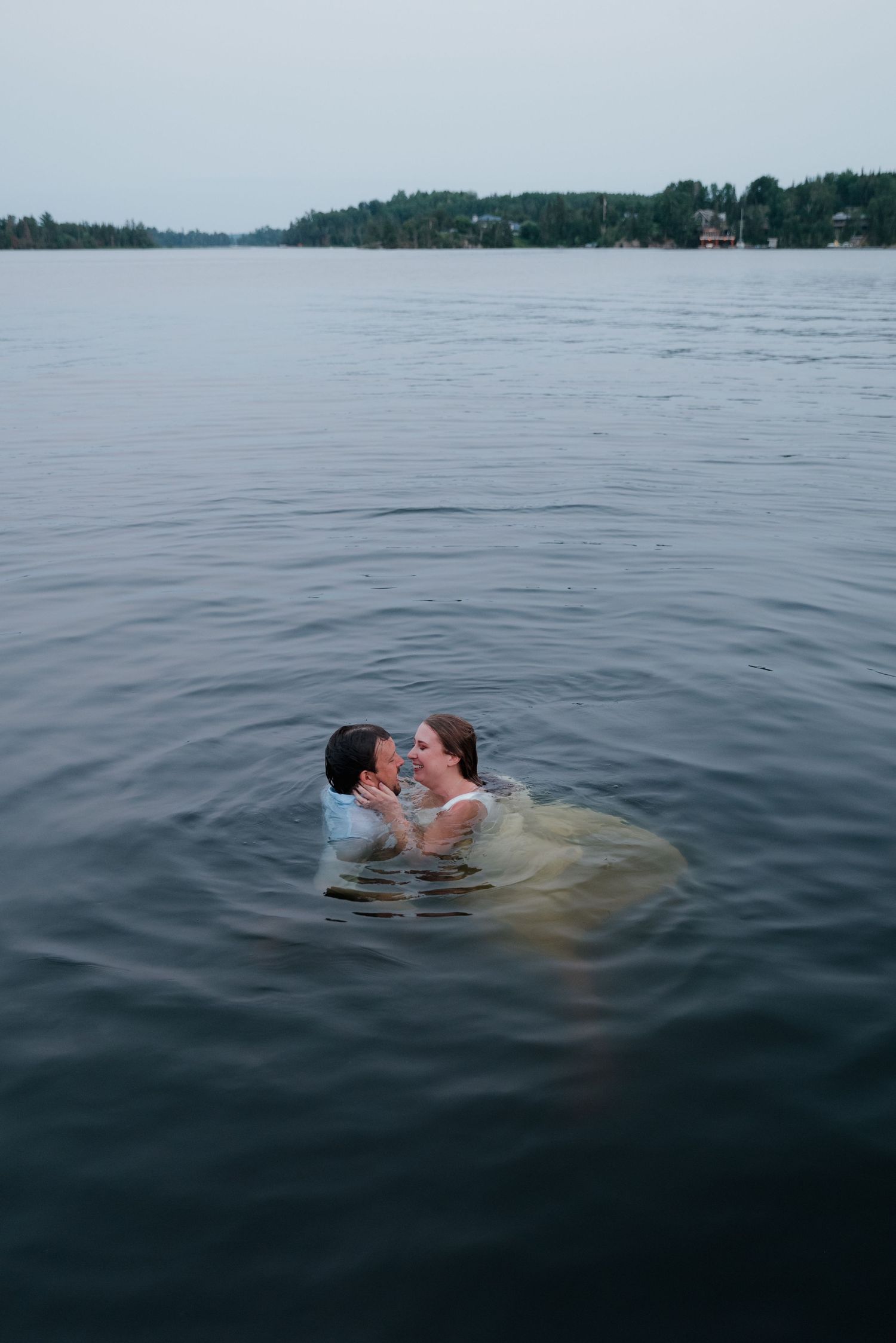 A couple embraces while swimming in a misty lake with forested shoreline visible in the background.