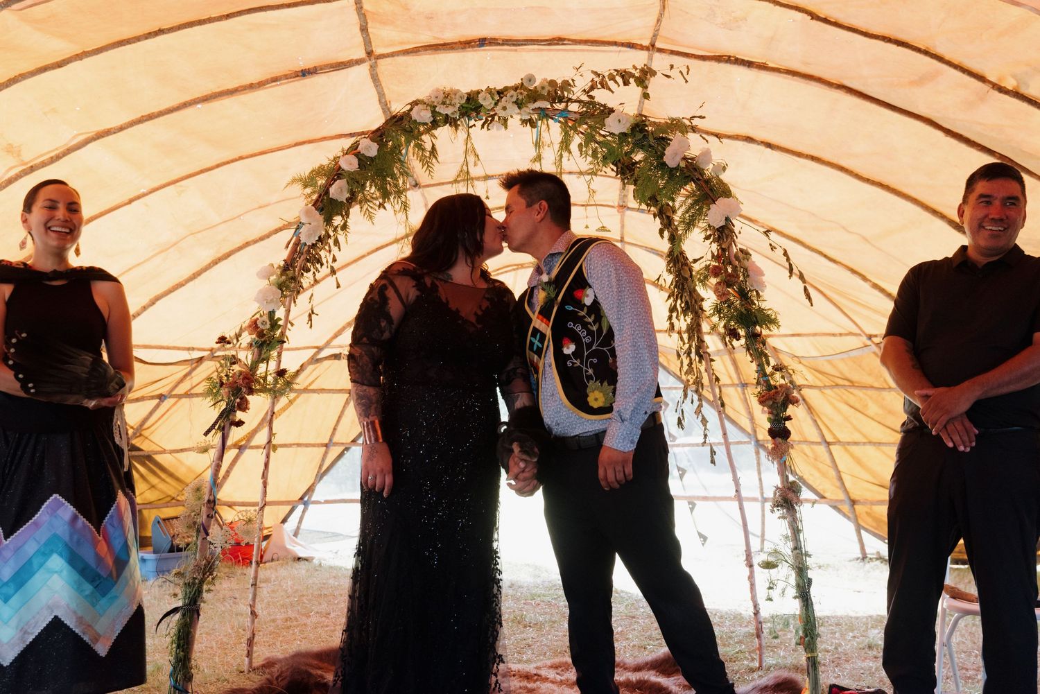 Couple shares first kiss under floral arch at Native American wedding ceremony with witnesses standing beside them.