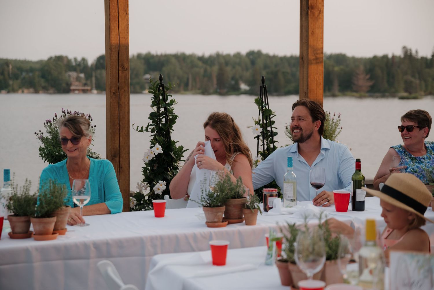 Wedding guests gather under a wooden pergola by a lake for an outdoor reception celebration.