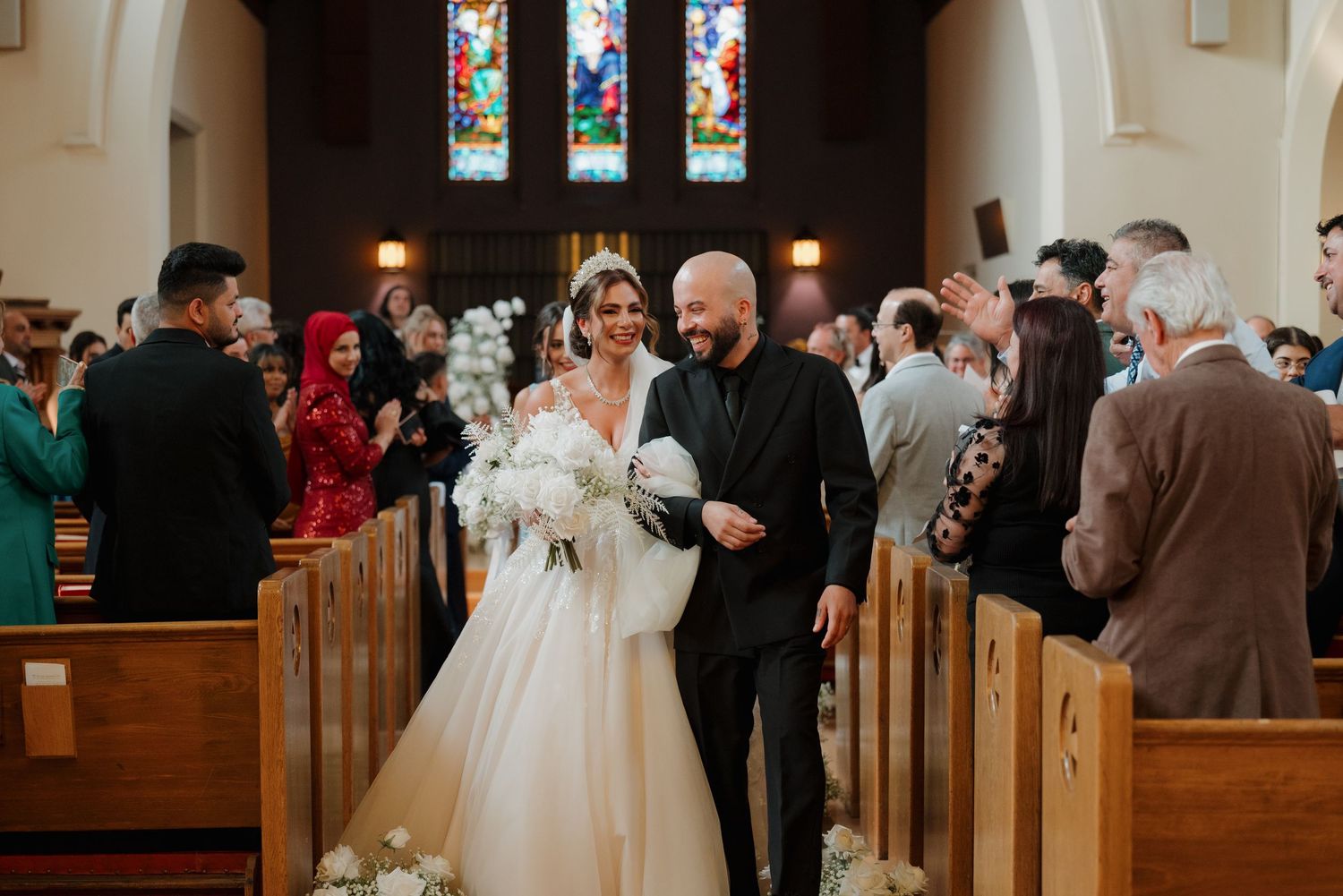 Wedding ceremony guests gather inside a traditional church with stained glass windows and wooden pews.