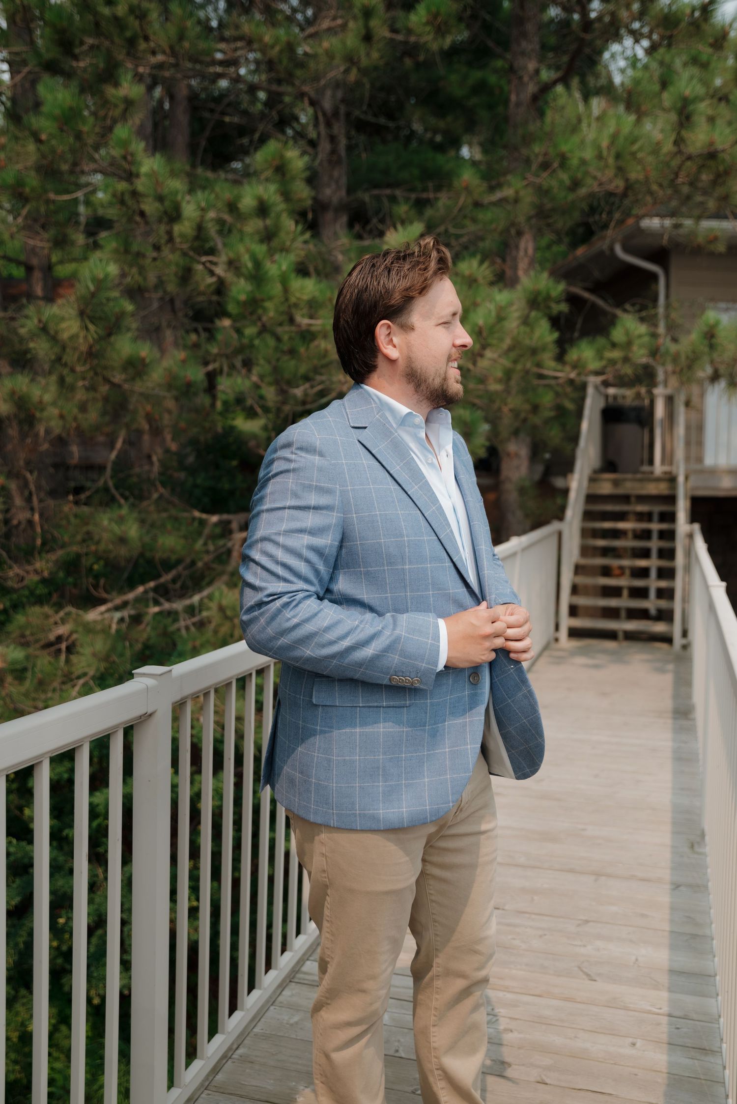 Person in light blue suit jacket and khaki pants stands on outdoor wooden deck surrounded by greenery.