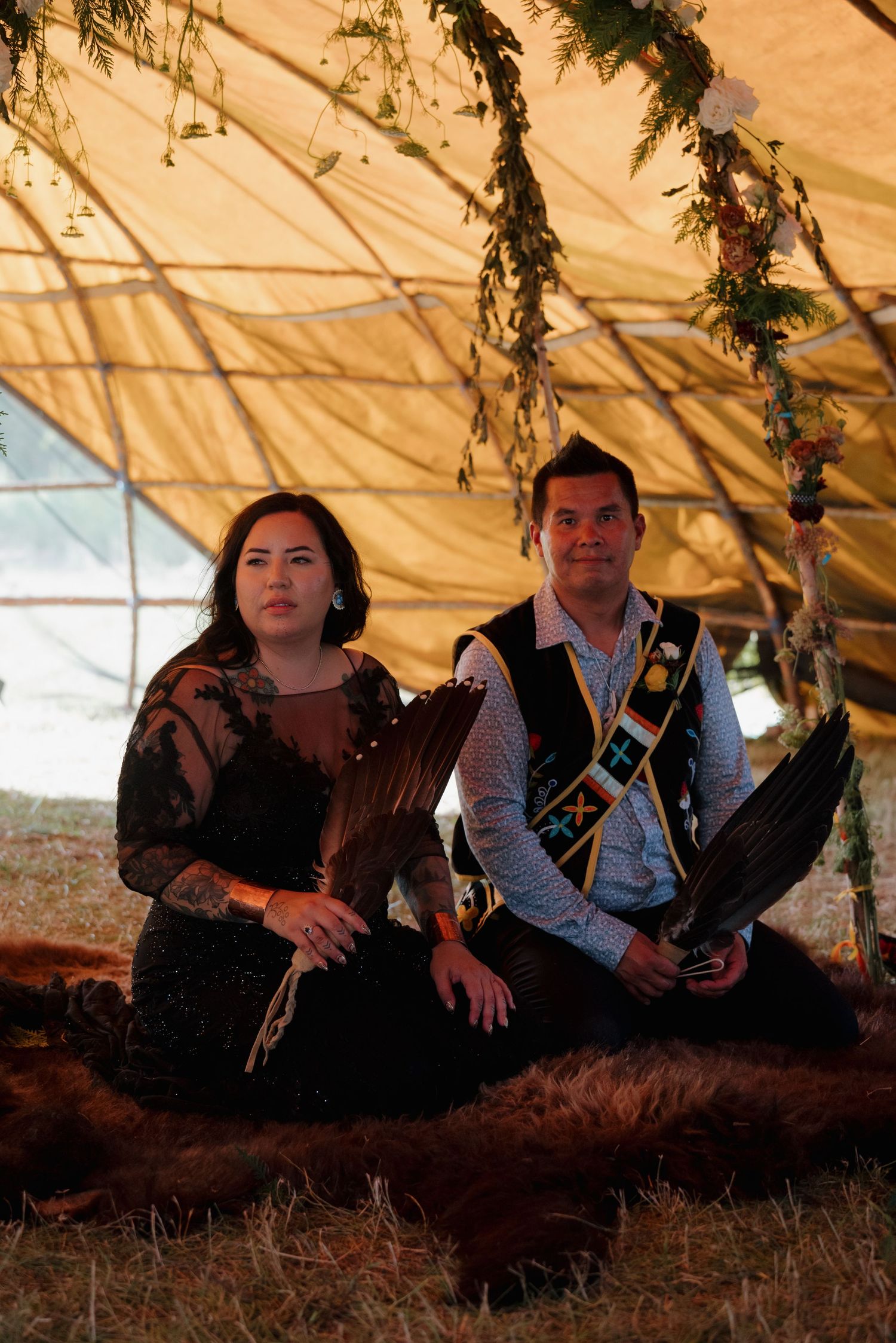 Native American couple in traditional ceremonial attire sitting on fur beneath a floral-adorned hide arbor during a wedding ceremony.