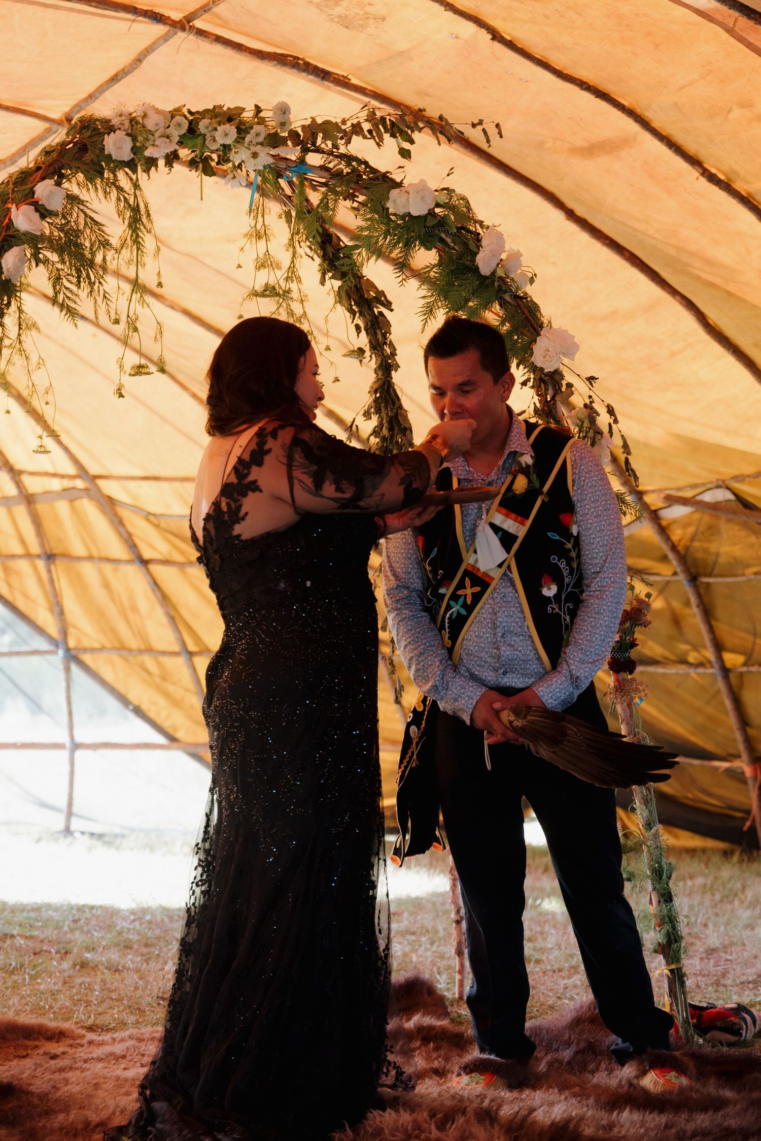 Couple exchanges vows under floral arch inside ceremonial tent, groom wearing Indigenous regalia sash at wedding ceremony.