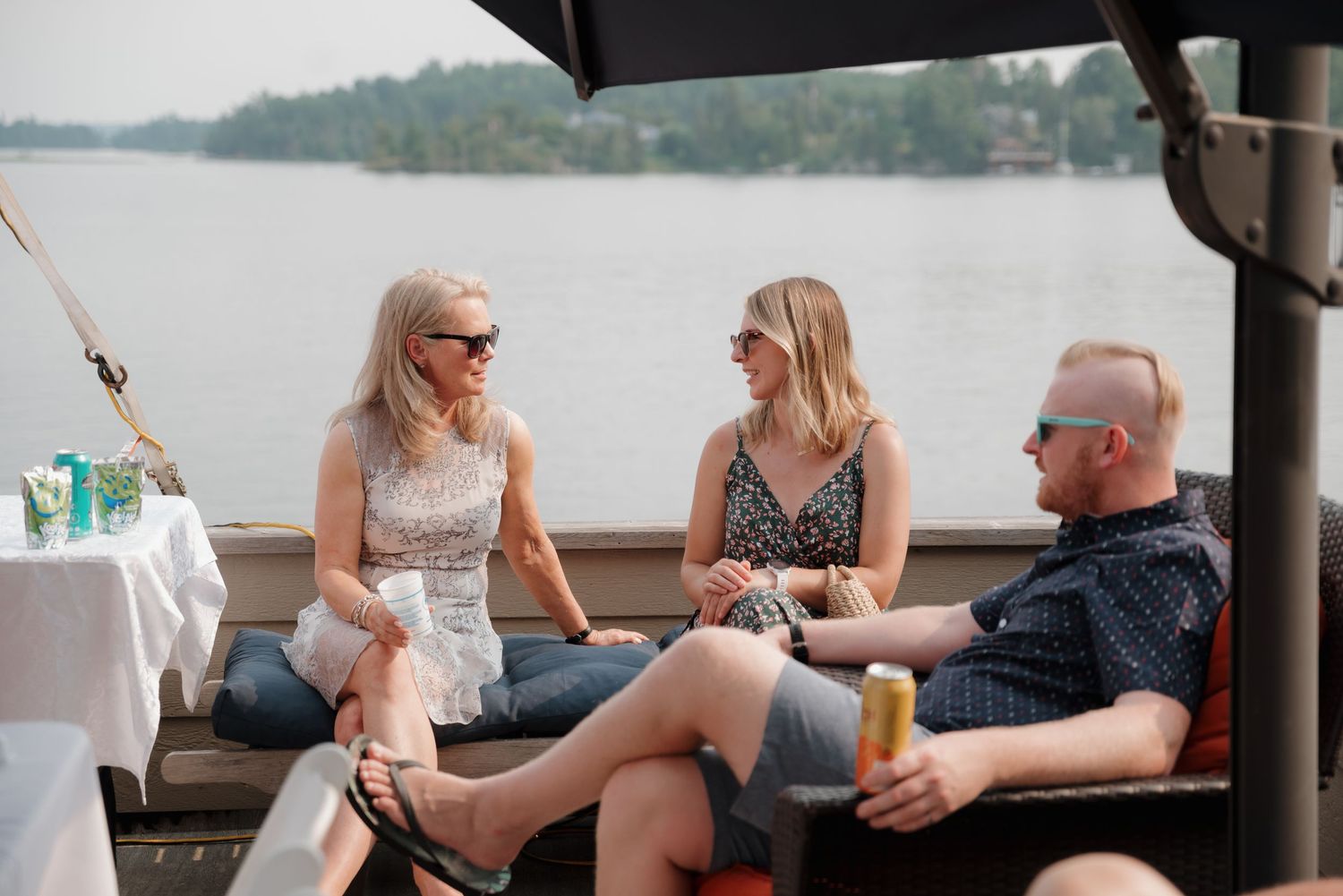 People relaxing and socializing on a covered deck overlooking a scenic lake on a summer day.