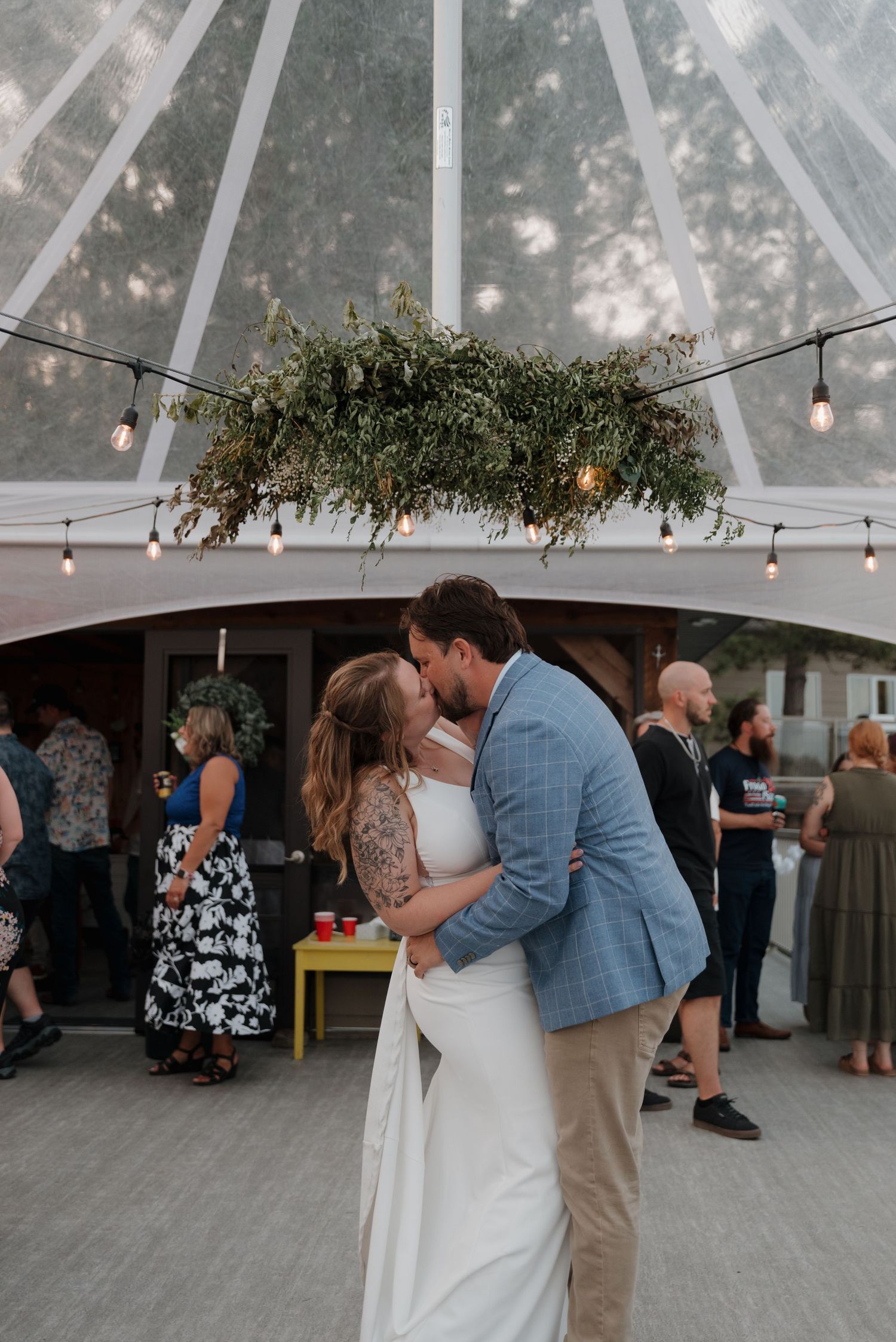 Wedding reception under a clear tent with string lights and hanging greenery decorations.