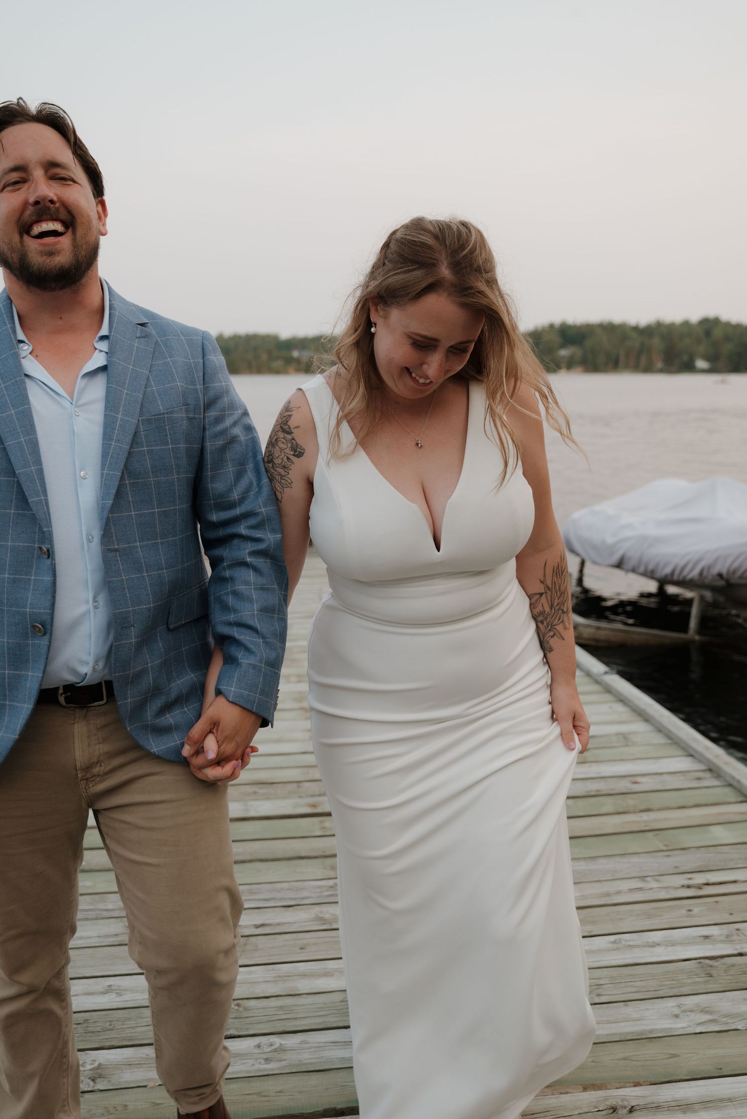 A couple in wedding attire shares a joyful moment on a wooden dock by the water.