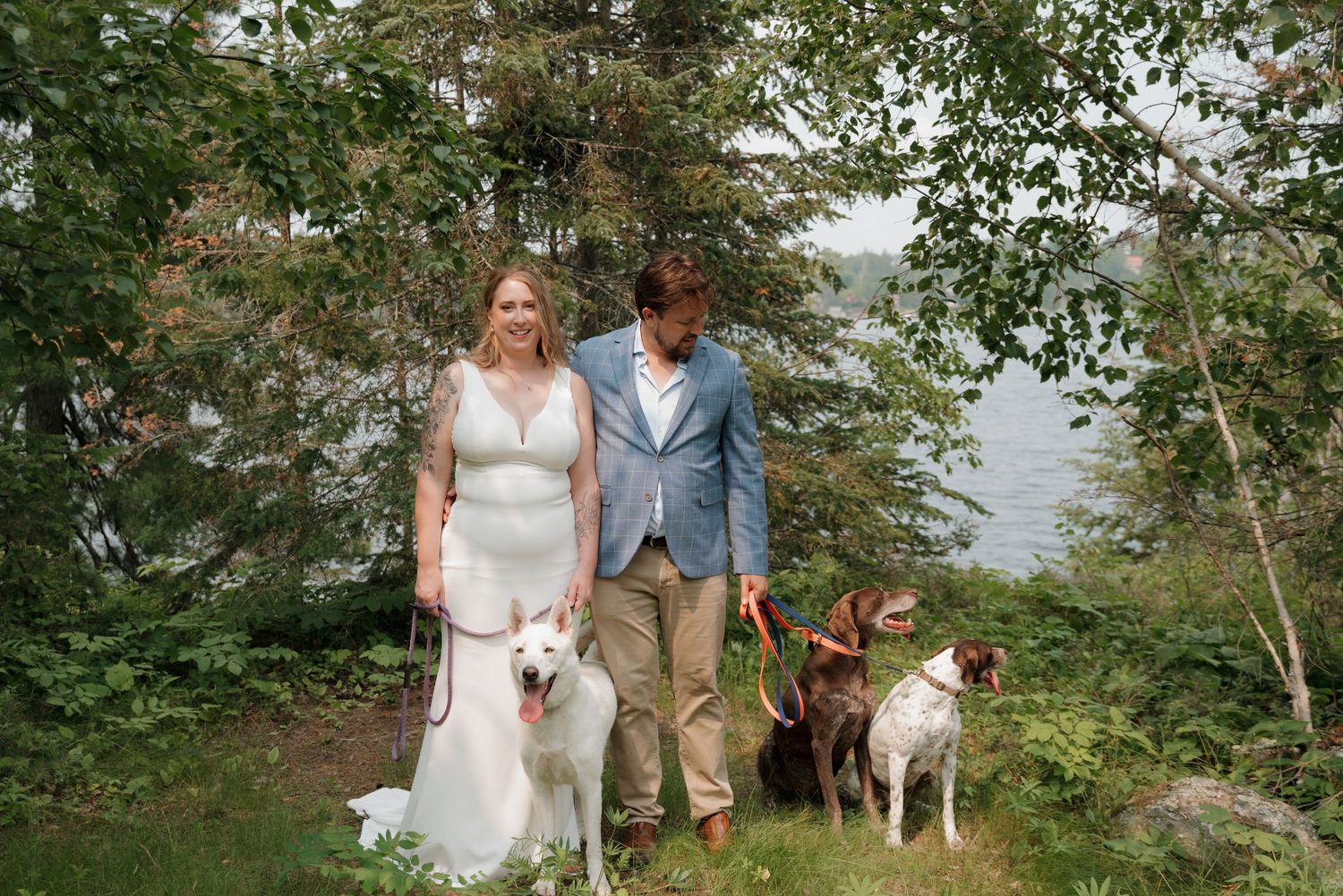 A couple and their dog pose together in a scenic outdoor setting surrounded by lush greenery and trees.