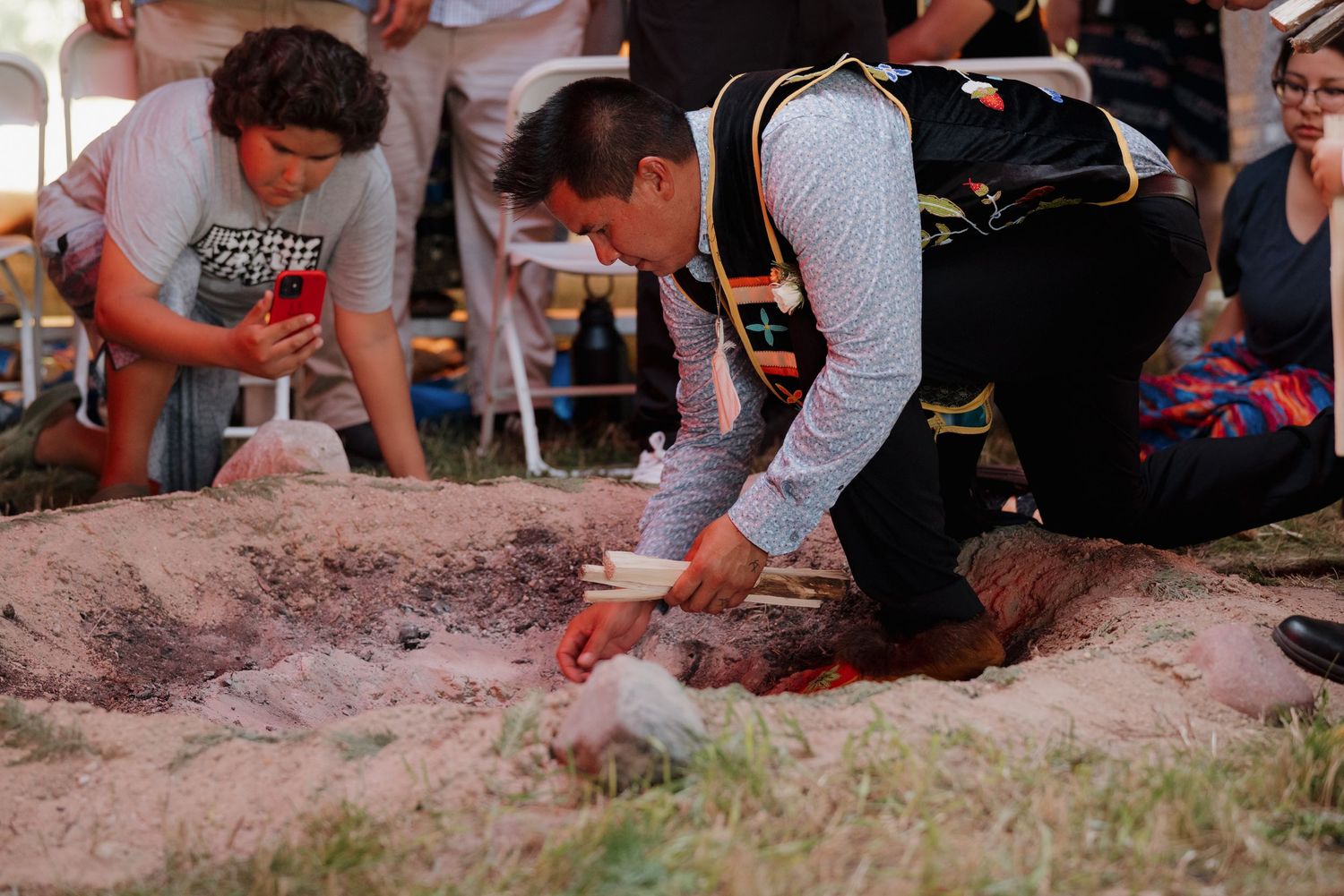 A man in traditional Native American attire kneels over a ceremonial sand pit during an indigenous cultural ceremony outdoors.