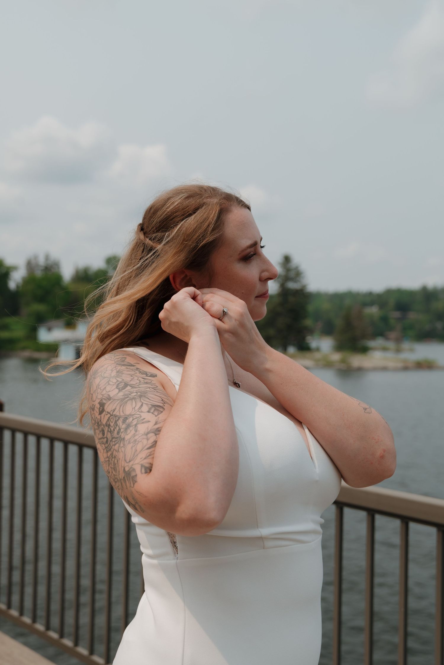 Woman in white dress with tattoos stands at waterfront railing during outdoor photoshoot.
