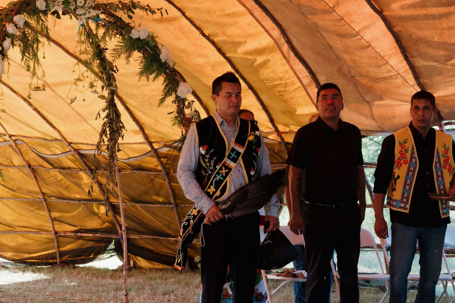 Three men in traditional cultural attire stand under a large golden tent decorated with greenery.