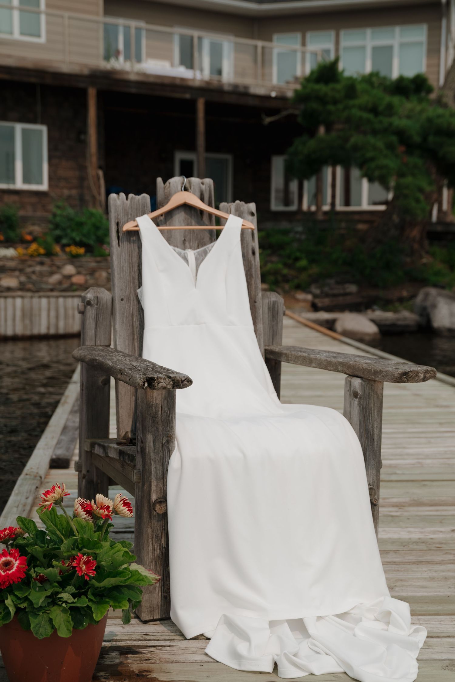 White wedding dress displayed on wooden dock with red flowers and lake in background.