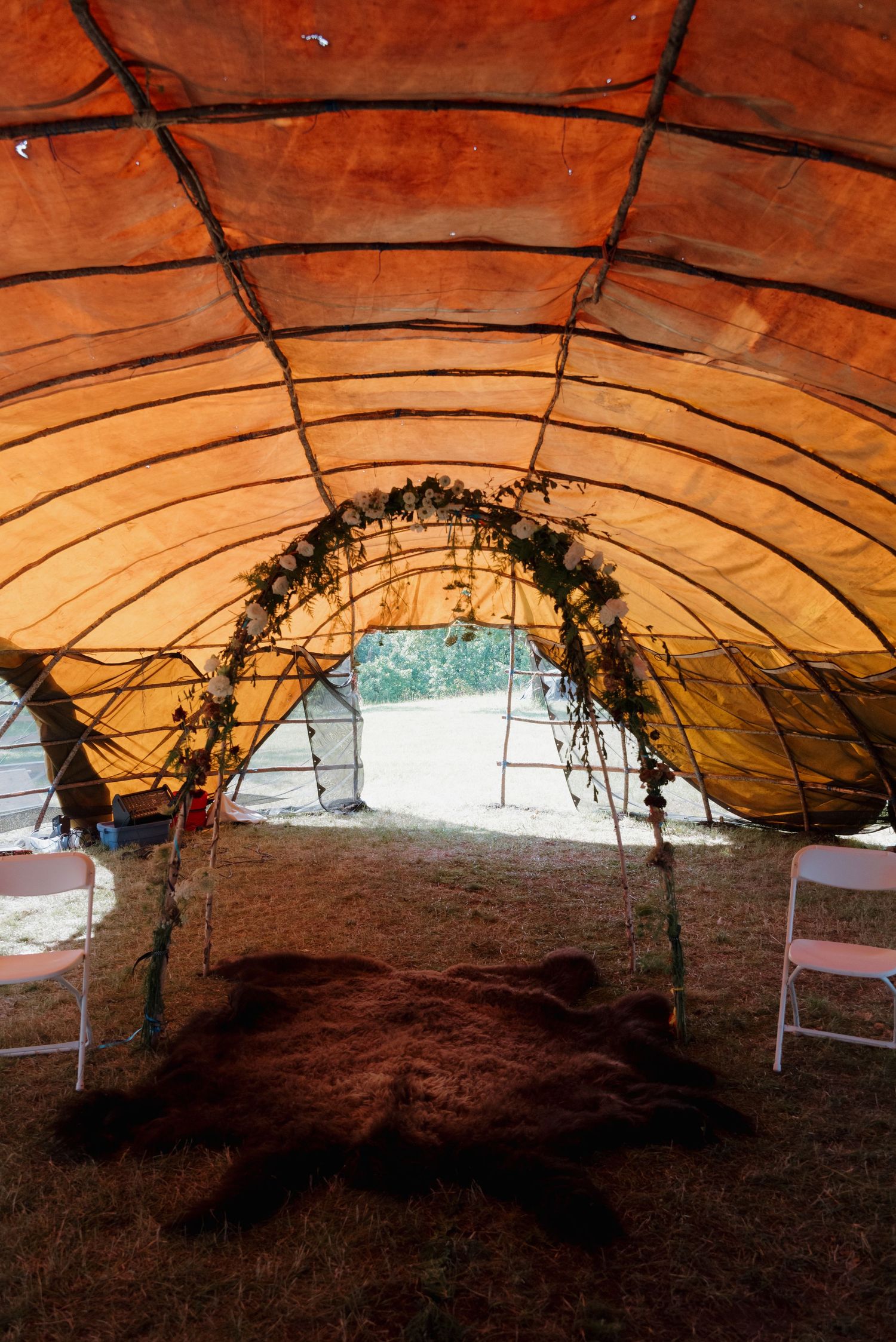 Interior of a Native American ceremonial arbor with orange hide covering, floral arch, animal fur rug, and white folding chairs.