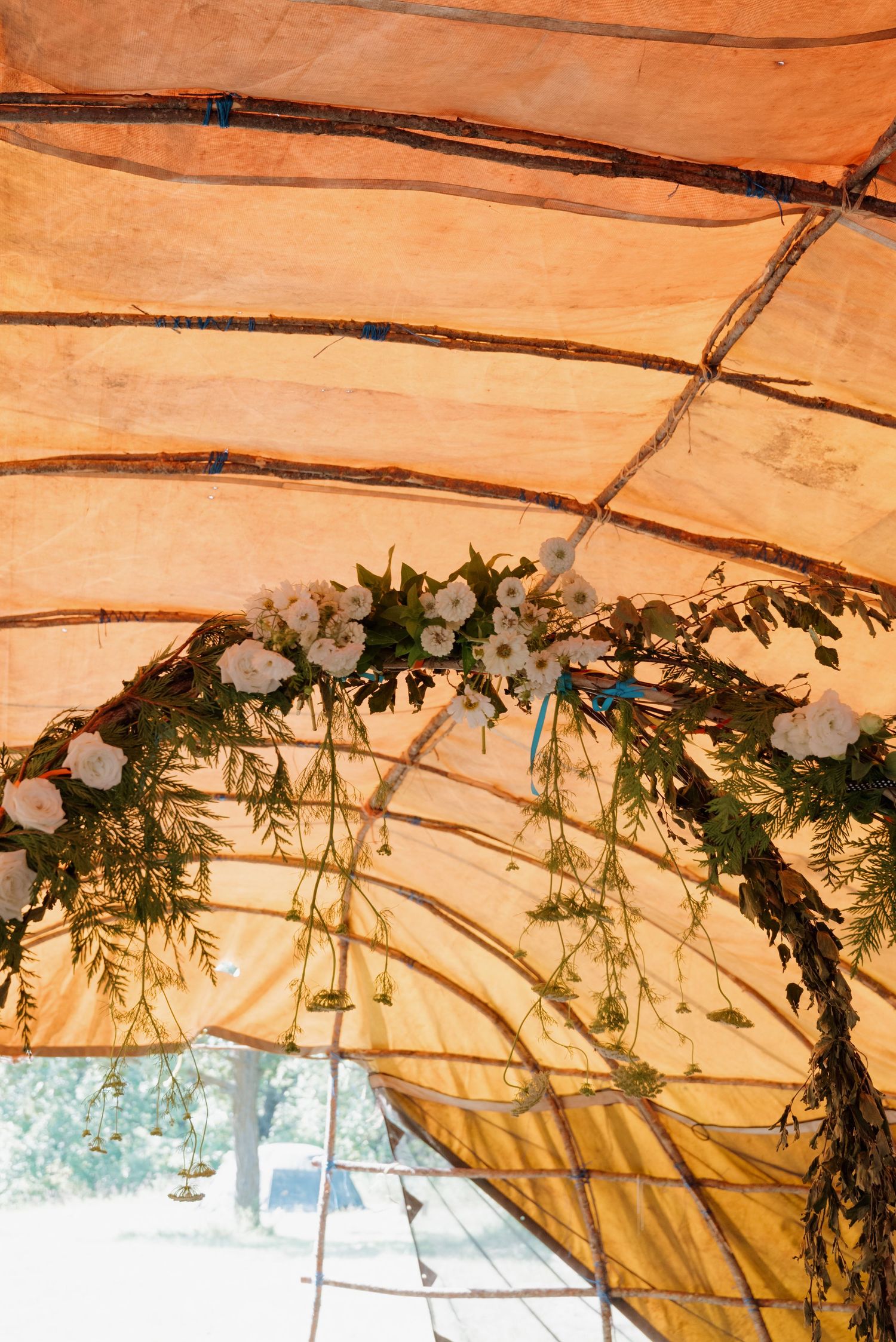 Floral arch with white roses and greenery hanging from a rustic wooden frame inside an orange canvas tent.