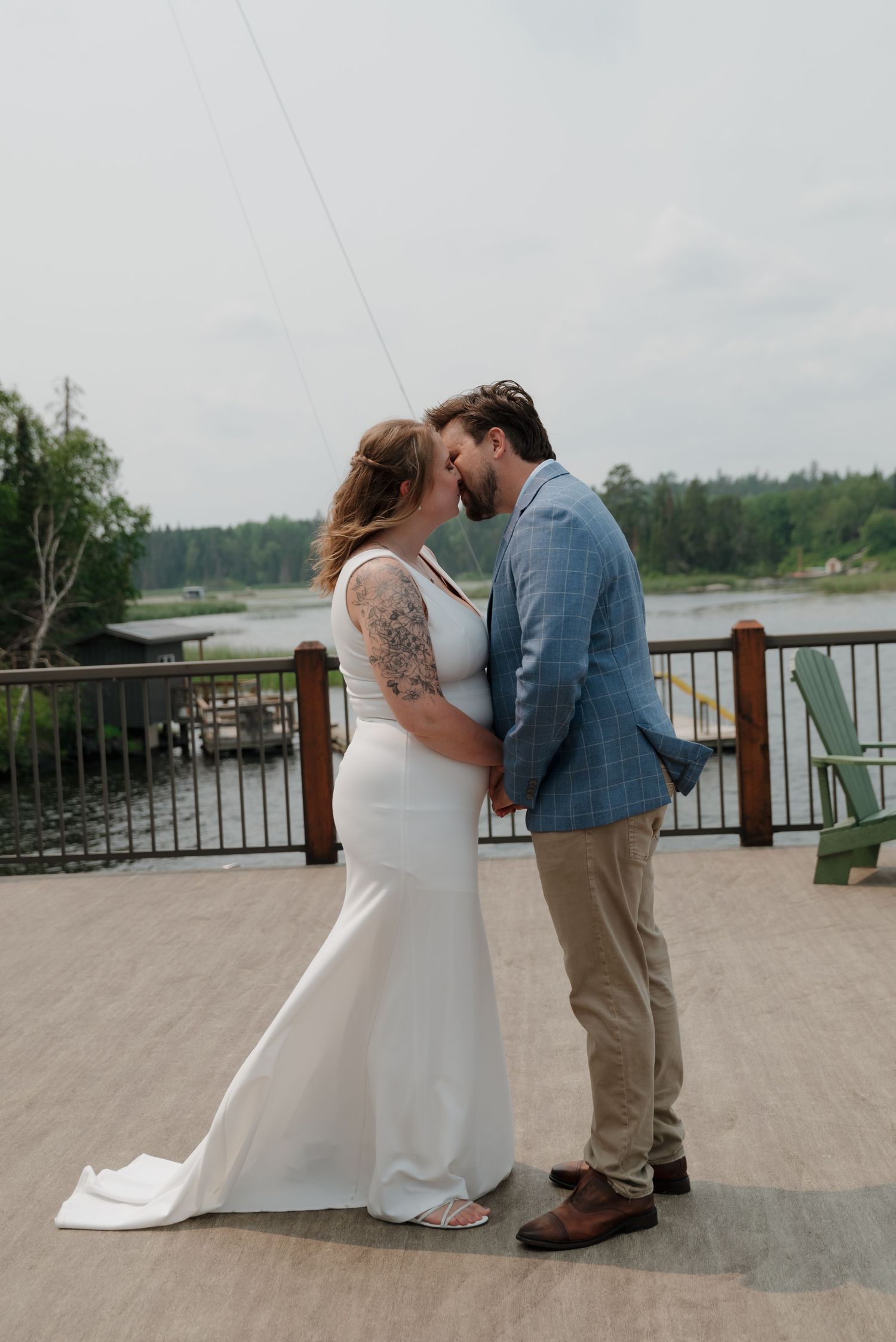 A romantic moment captured on a lakeside deck as a couple in wedding attire share an intimate embrace.