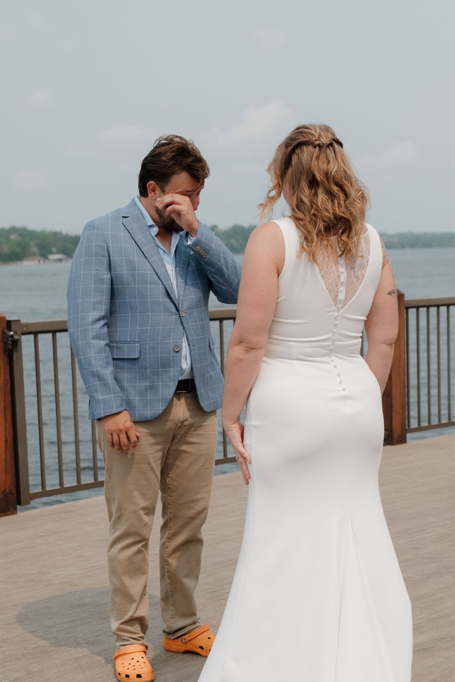 A couple in wedding attire share an intimate first look moment on a waterfront deck.