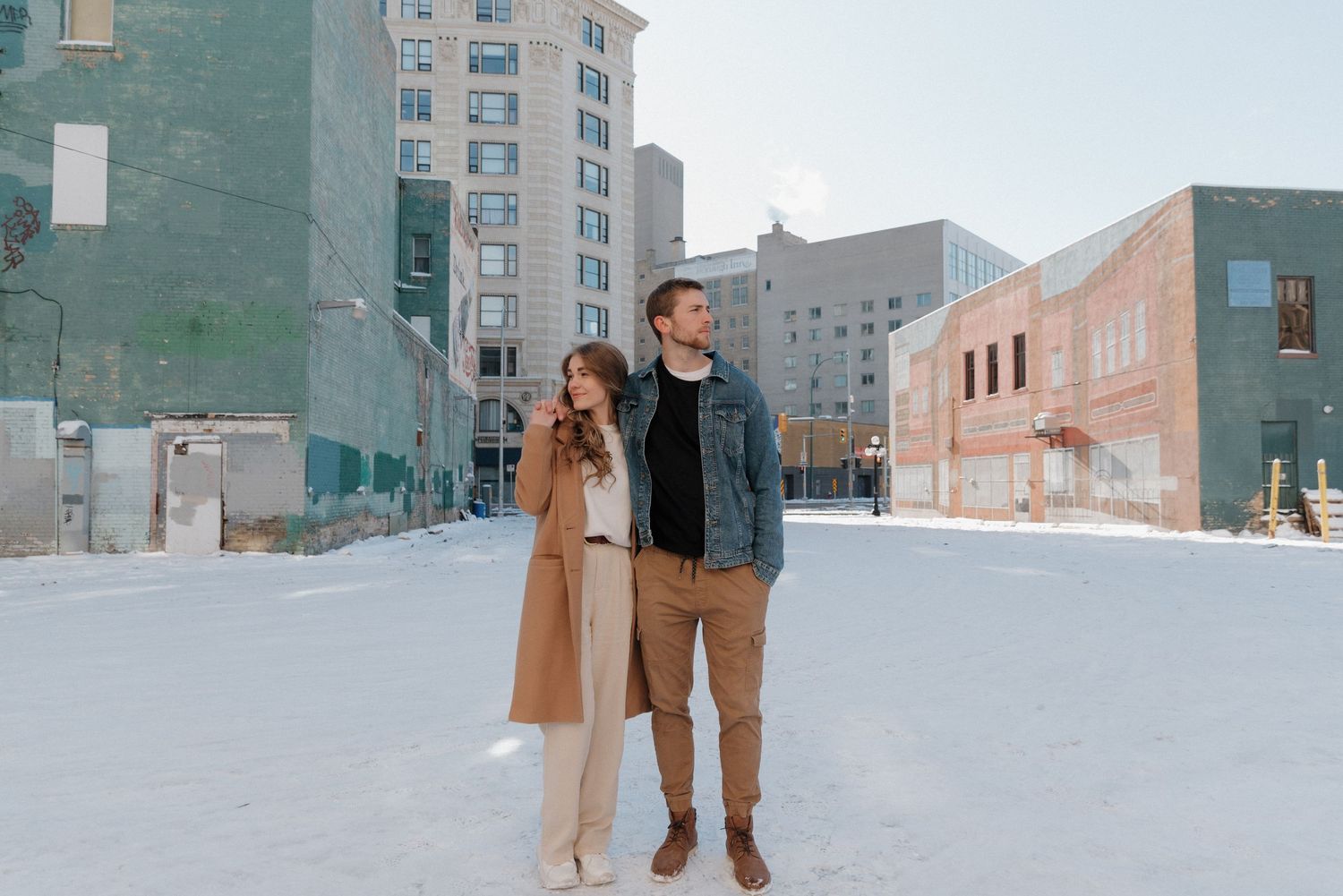 A couple poses together in a snowy urban setting with buildings in the background.