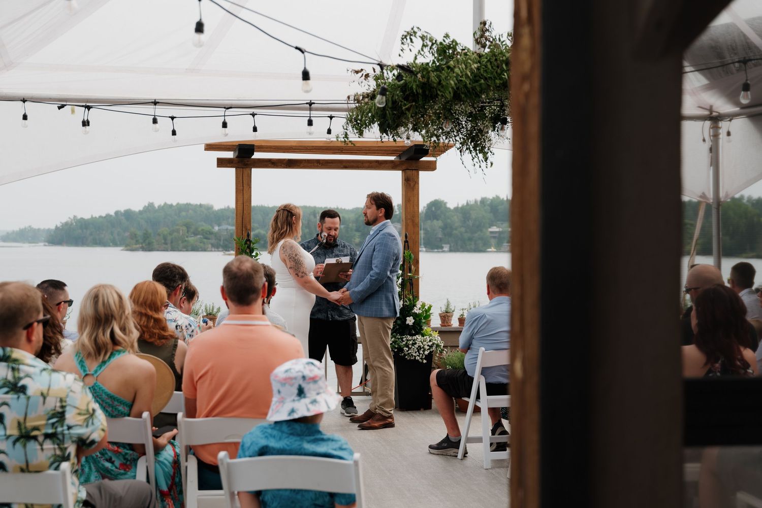 An outdoor wedding ceremony takes place under a wooden arch with string lights and waterfront views.