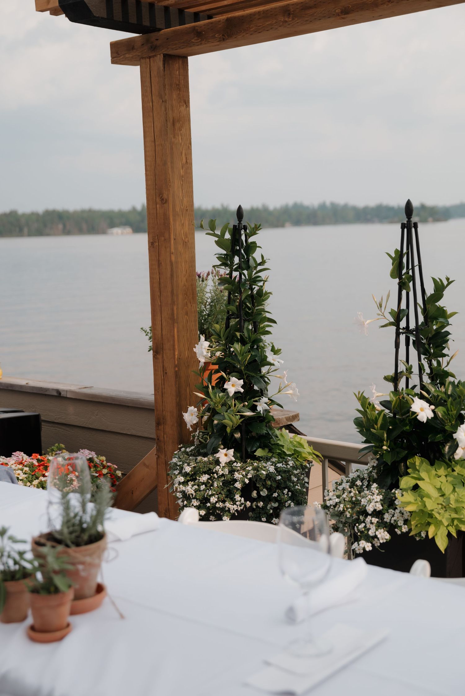 Elegant table setting with white linens and potted flowers overlooking a serene lake.