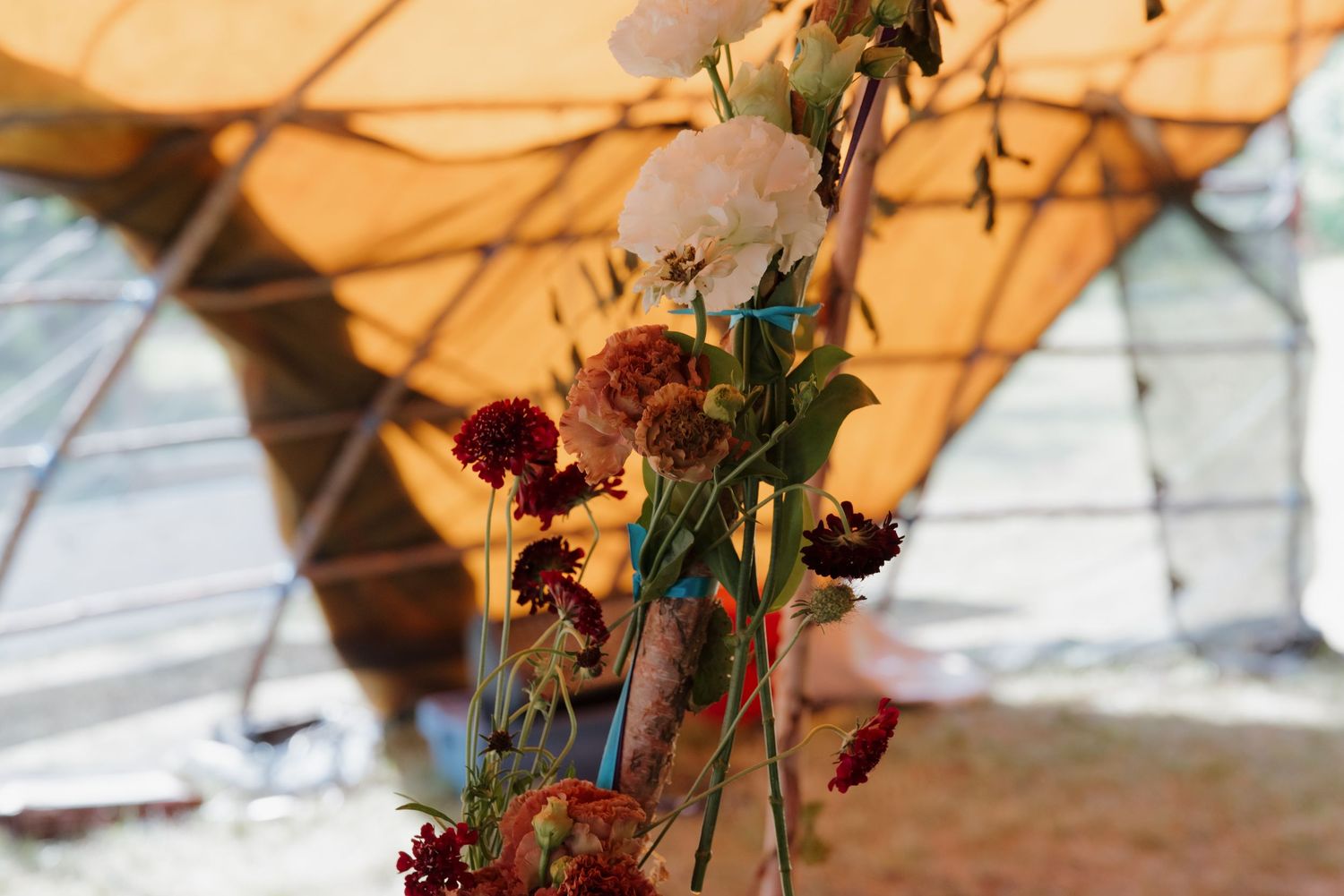 Rustic floral arrangement with red and white blooms against a warm orange tent backdrop outdoors.
