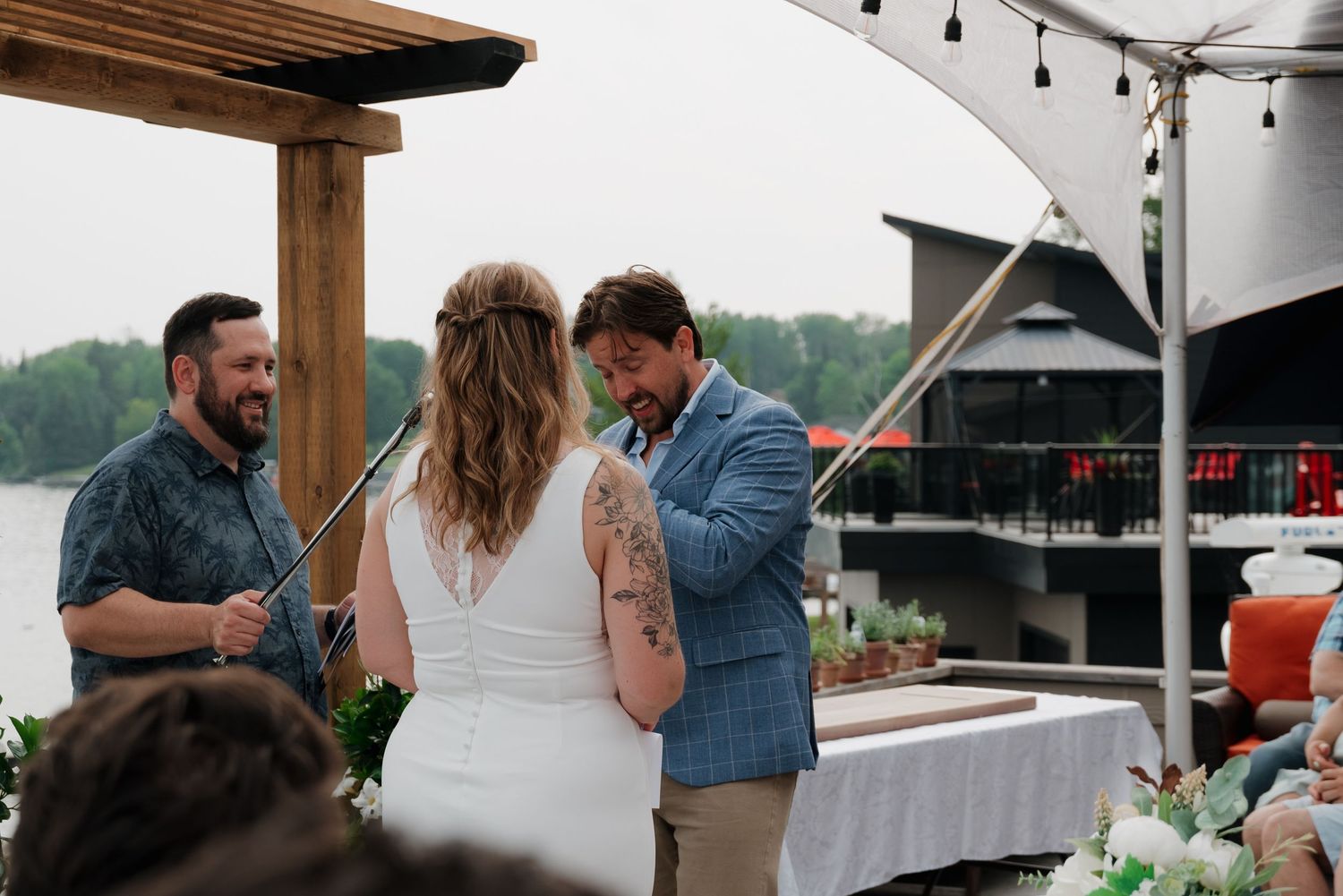 Guests mingle under wooden beams and string lights at an outdoor lakeside venue.