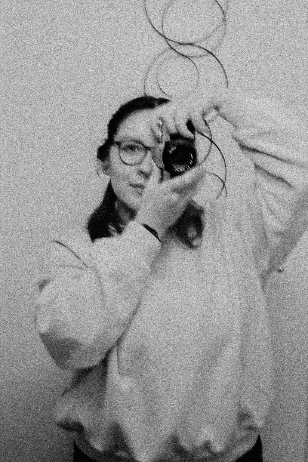 Woman in white sweatshirt and glasses holding camera up to take photo, black and white.