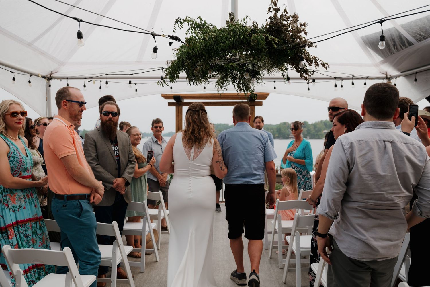 Wedding reception guests mingle under a white tent with string lights and waterfront views.