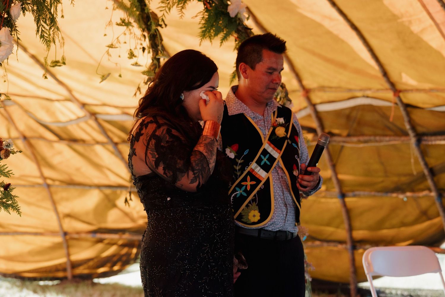 Emotional bride wipes tears while groom holds eagle feather during heartfelt Native American wedding ceremony under floral arch.