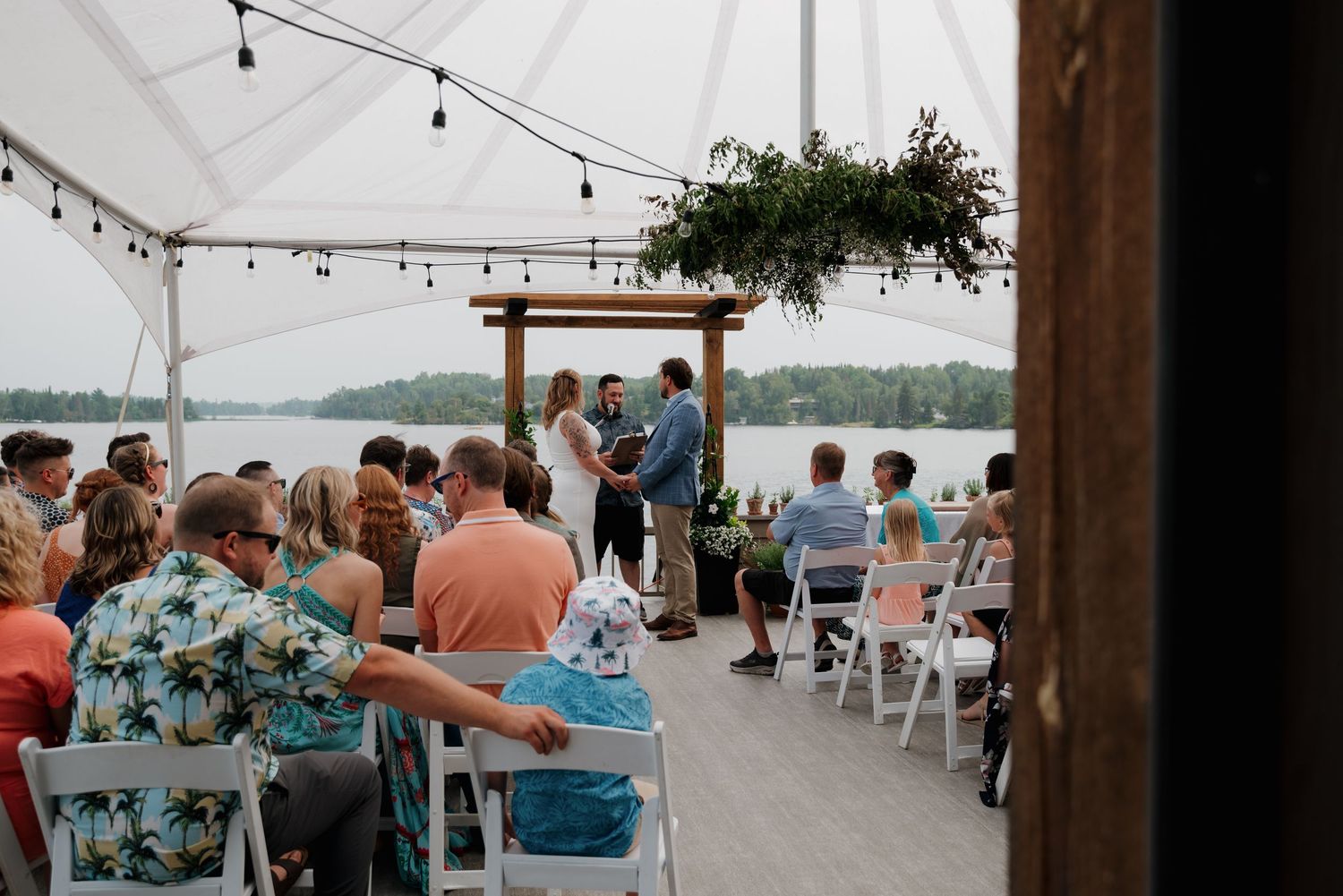 A rustic waterfront wedding reception venue with white chairs and blue table decor under string lights.