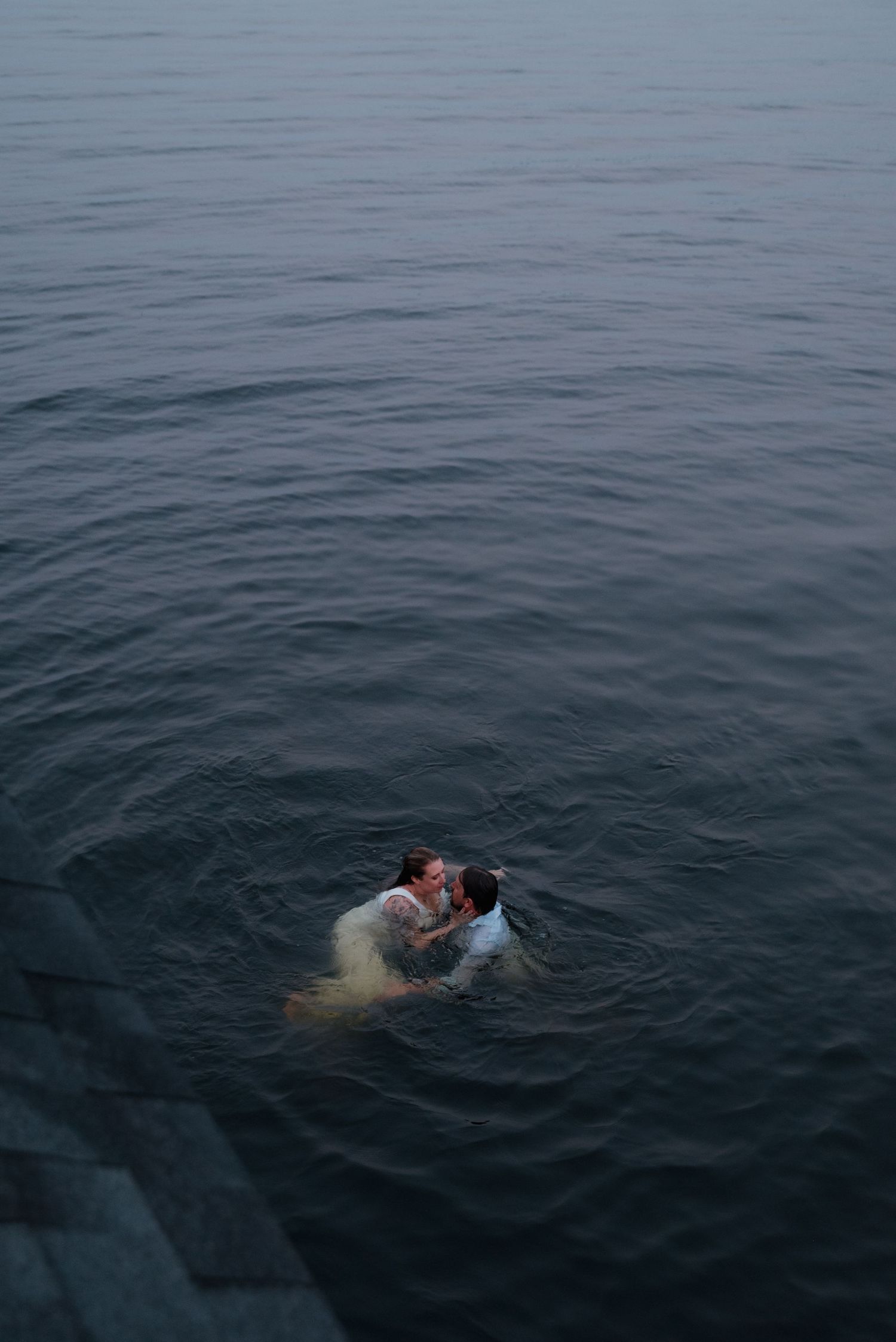 Two people share a romantic moment while swimming in dark waters at dusk.