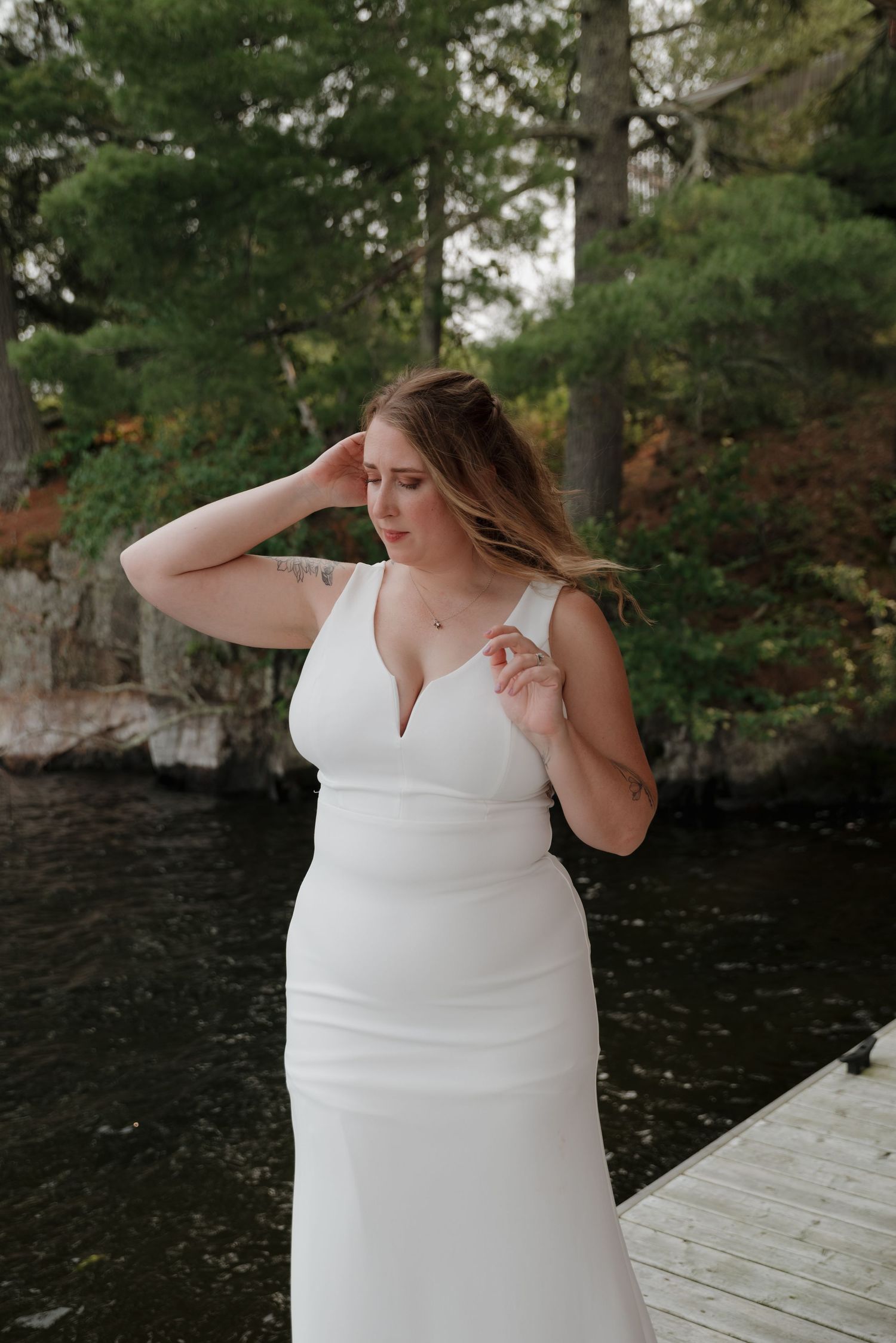 A person in a white wedding dress poses elegantly on a wooden dock by a lake surrounded by trees.