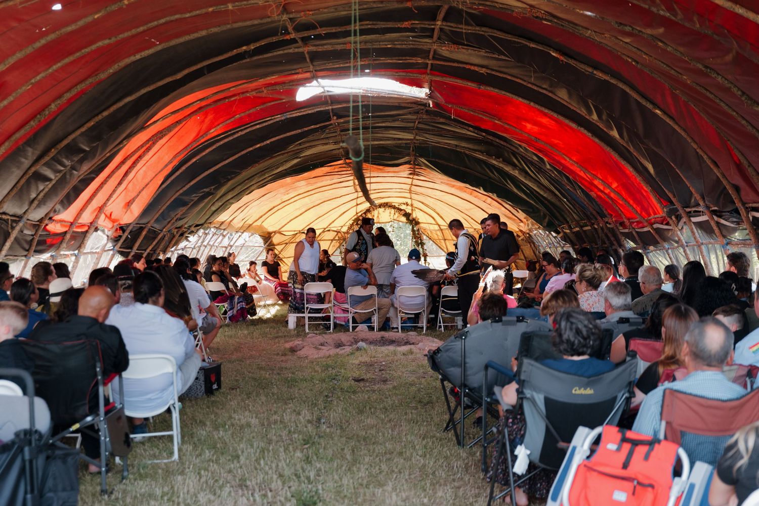 Large gathering of people seated inside a red-lit tent structure during an outdoor ceremony.