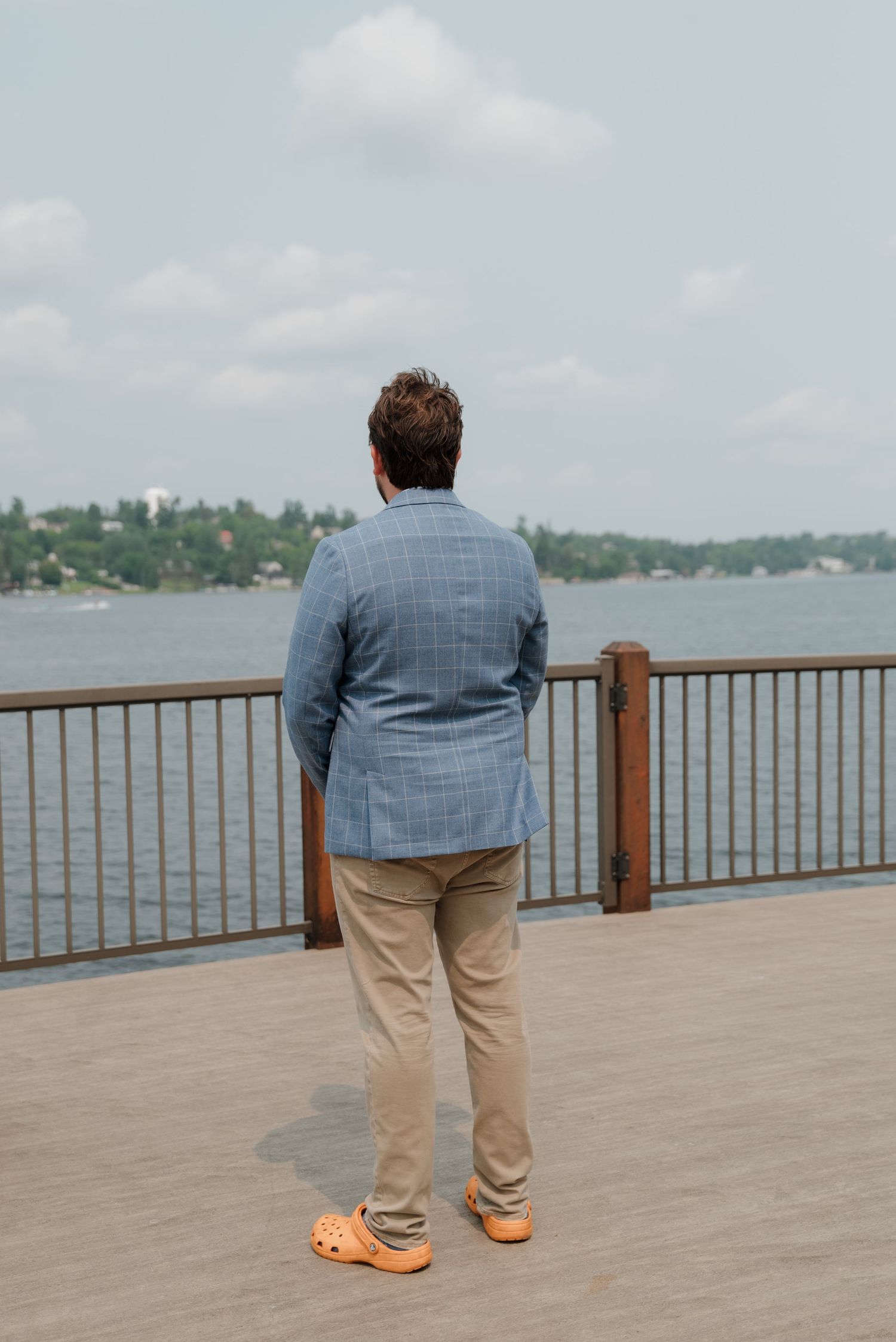 A figure in a blue blazer and khaki pants stands at a waterfront deck railing overlooking a scenic lake view.