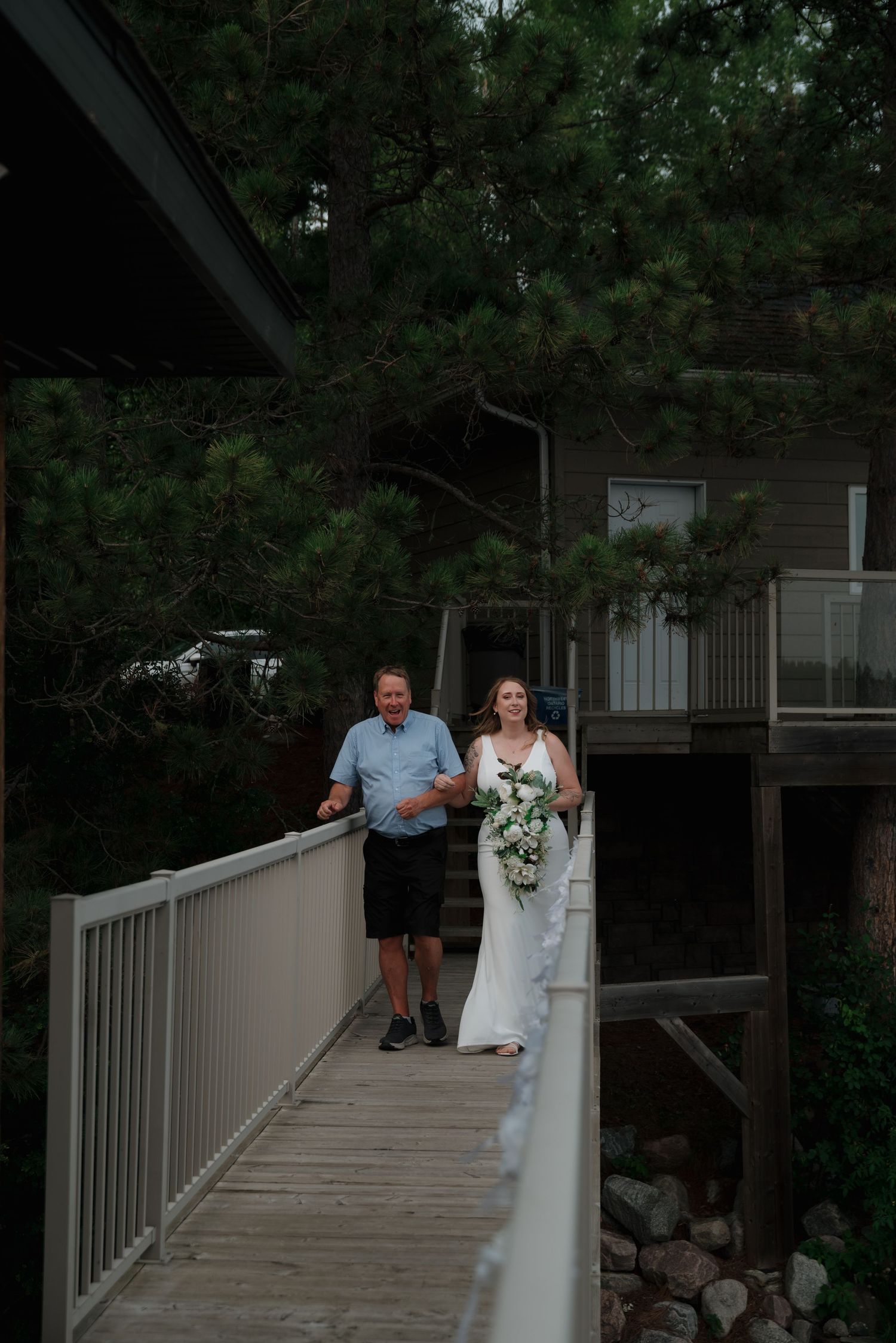 A wedding couple walks along a wooden boardwalk surrounded by trees at a lakeside venue.