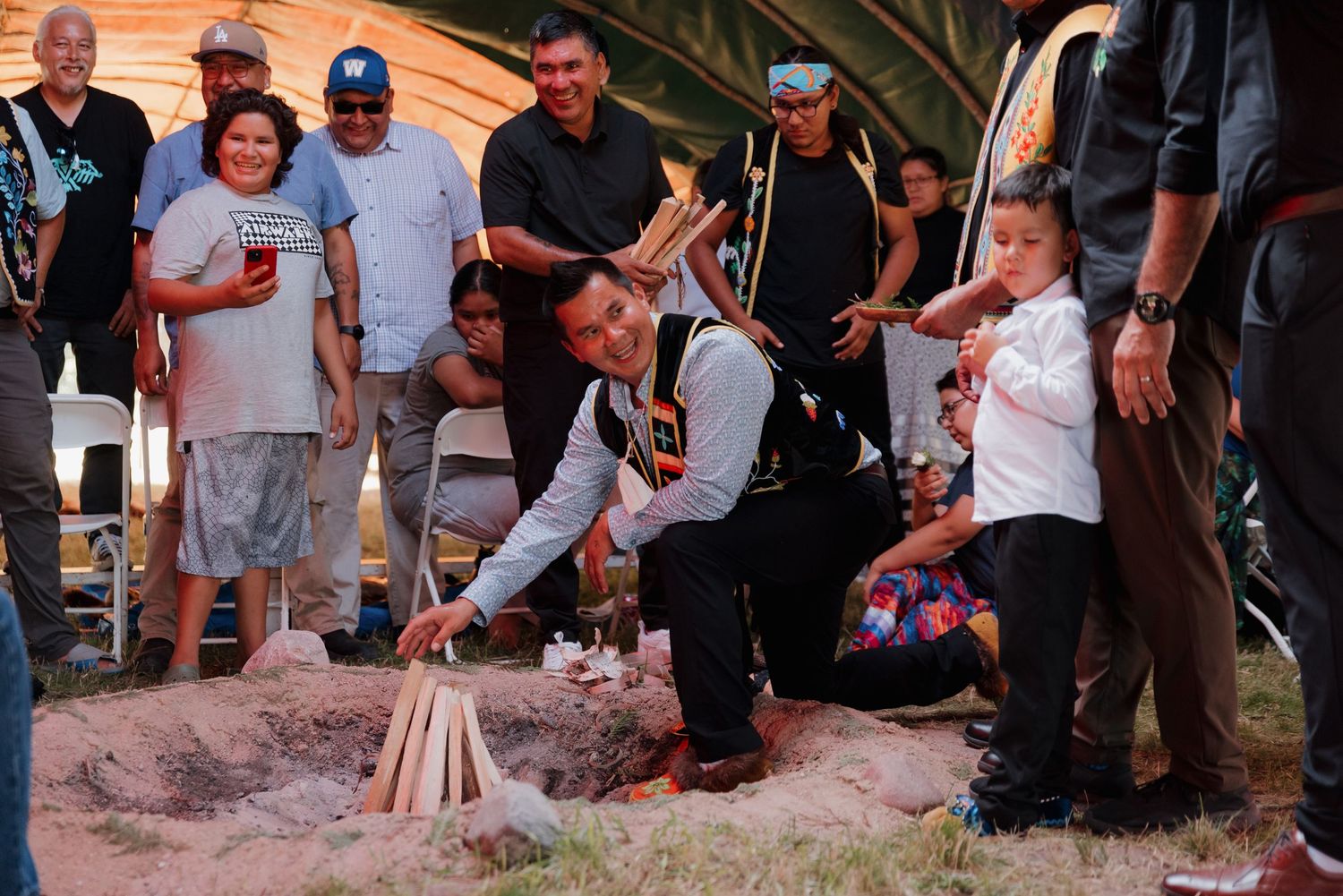 Native American elder kneeling to light a ceremonial fire surrounded by community members in traditional regalia indoors.