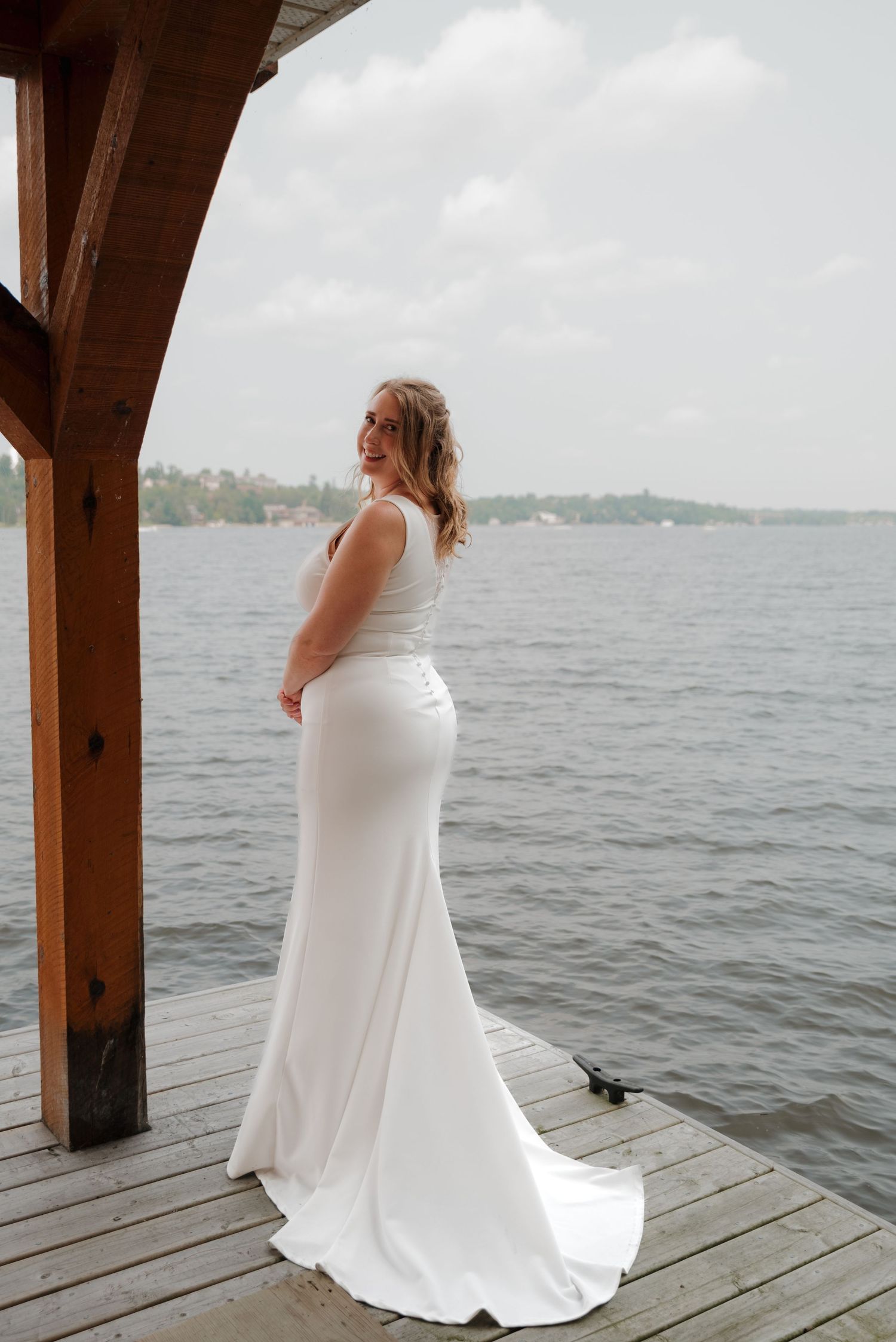 A bride in a flowing white dress stands on a lakeside dock during an overcast day.