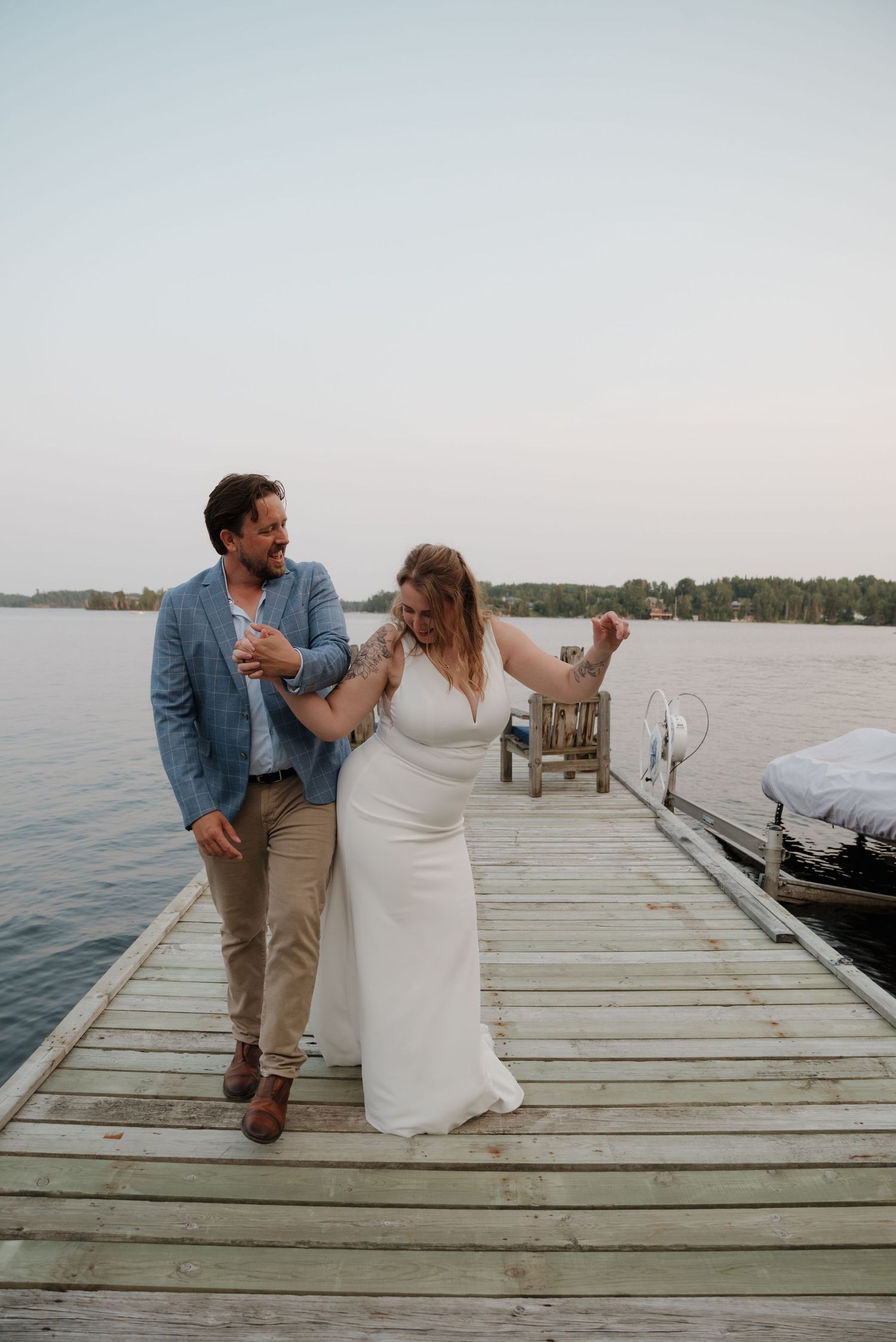 A couple shares a playful moment on a wooden dock by the water during a sunset photoshoot.