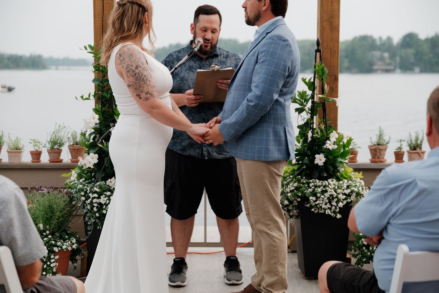 An outdoor lakeside wedding ceremony taking place on a decorated deck with potted plants.