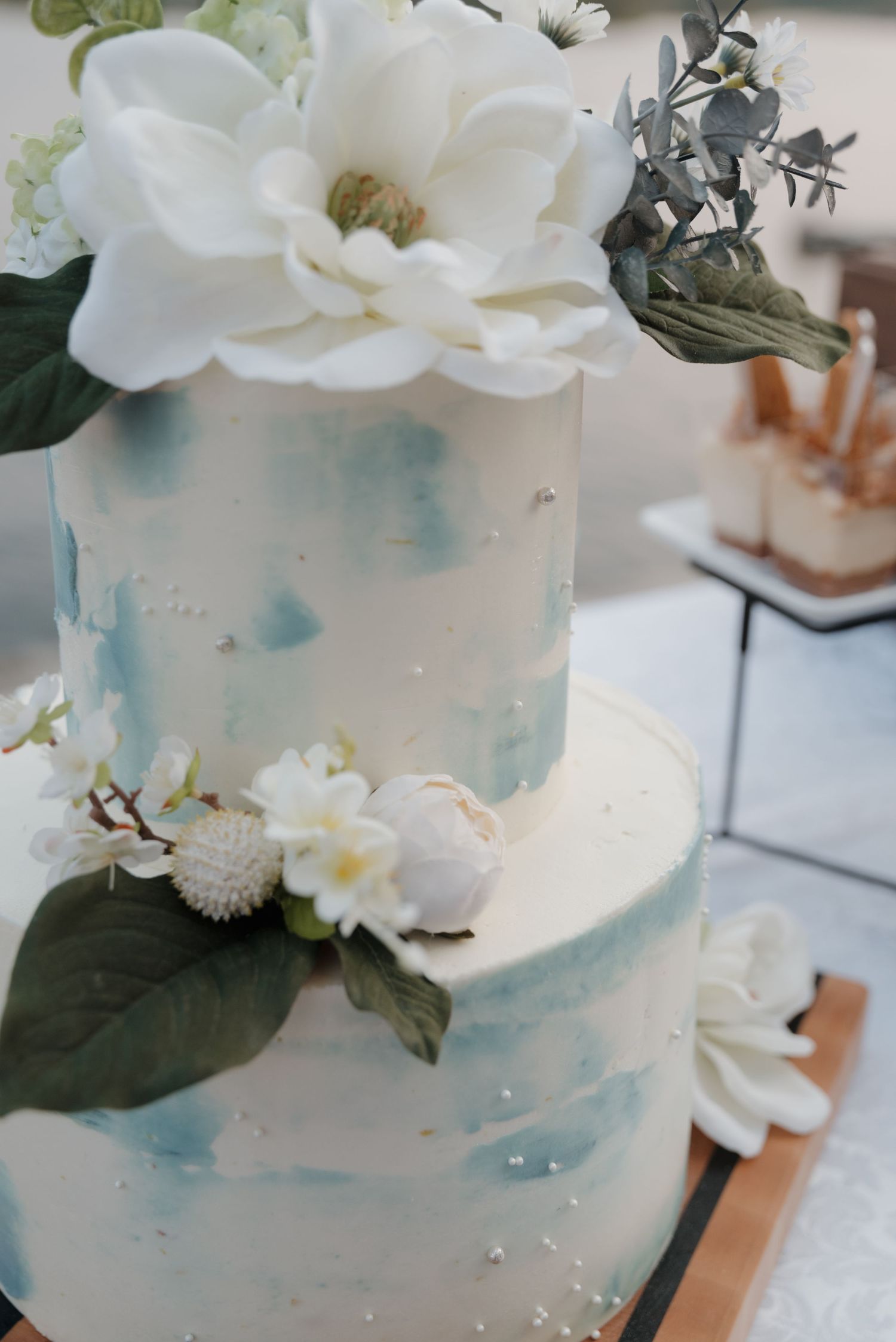 Elegant two-tier wedding cake with light blue watercolor design and white floral decorations on display.
