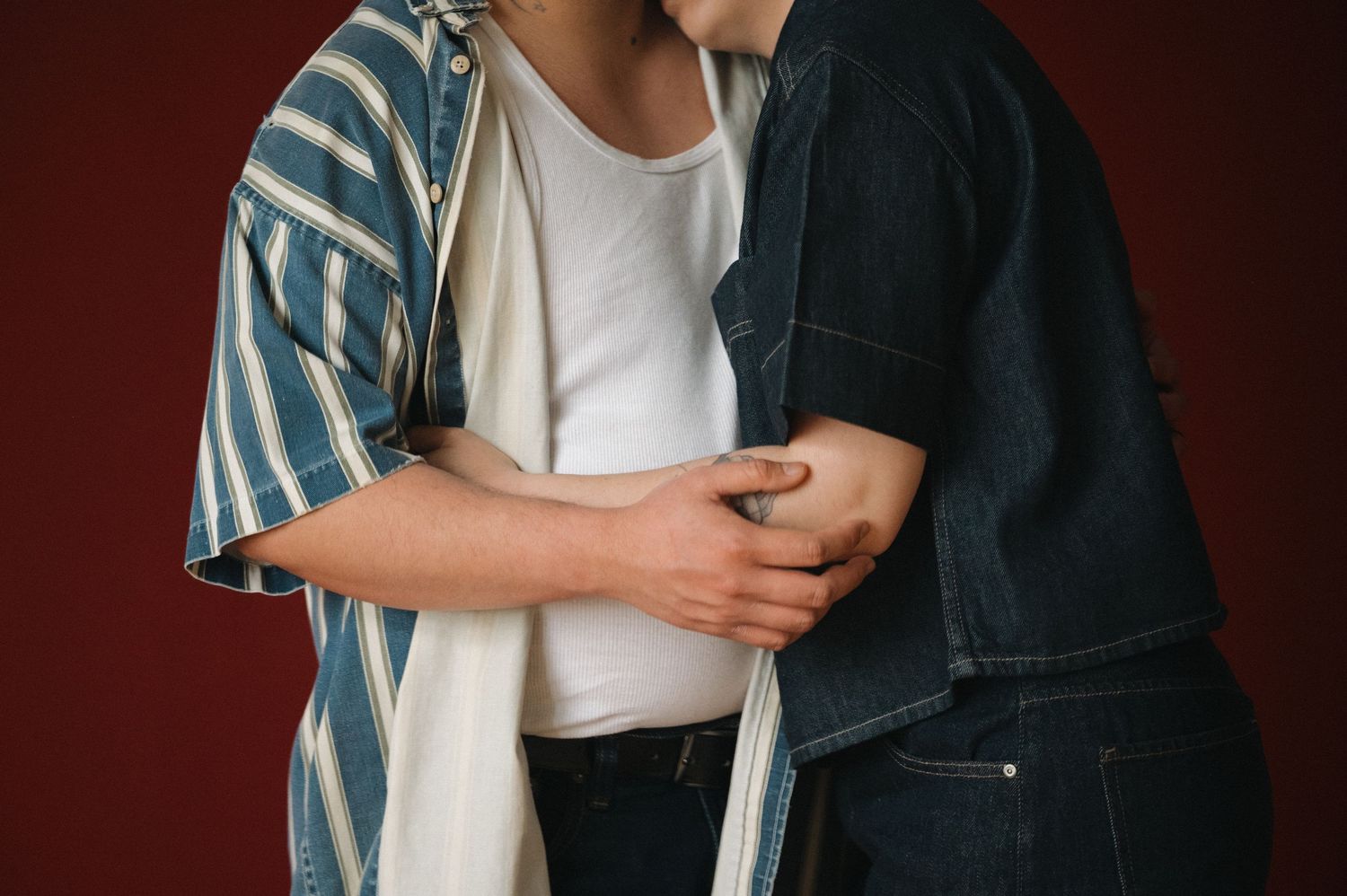 Close-up of two people embracing, wearing striped and denim shirts against a dark red background.