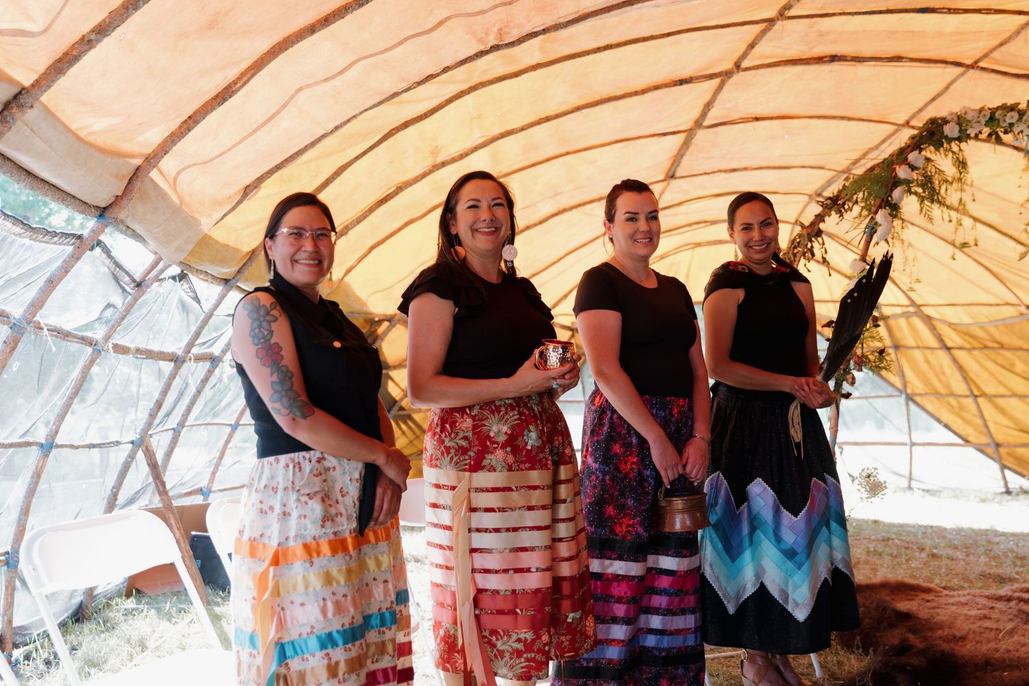 Four women in colorful traditional skirts stand smiling inside a tent-like structure during a cultural gathering.
