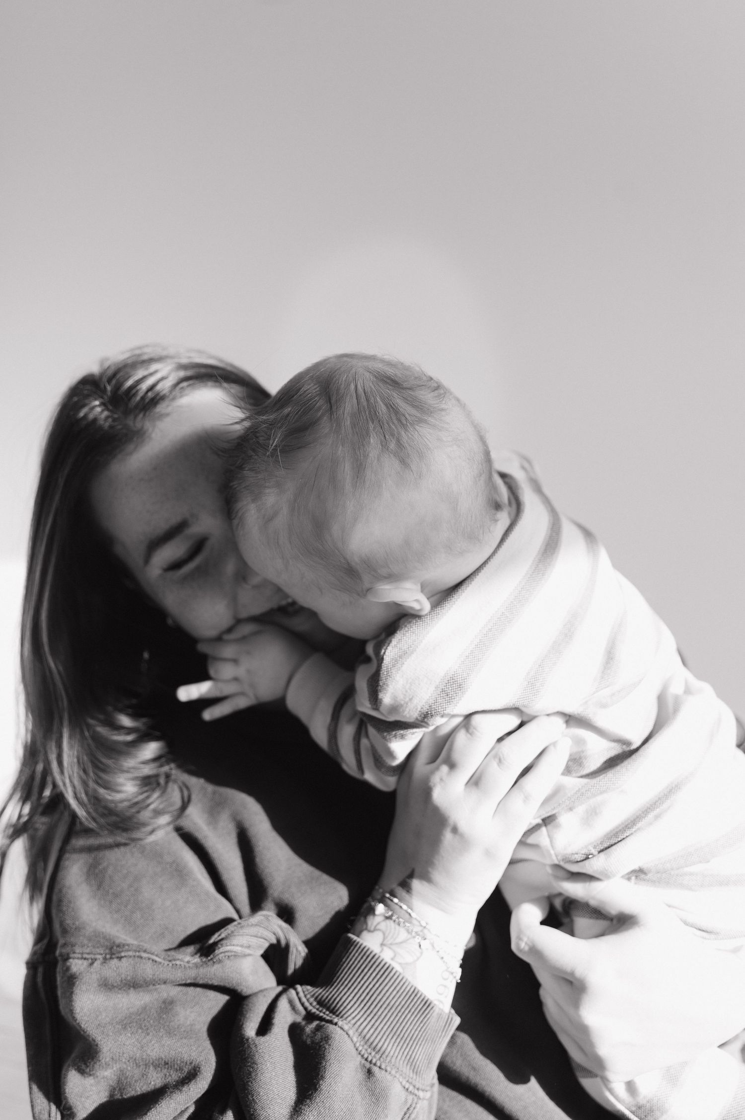 Black and white photo of a young mother tenderly kissing her baby against a bright background.