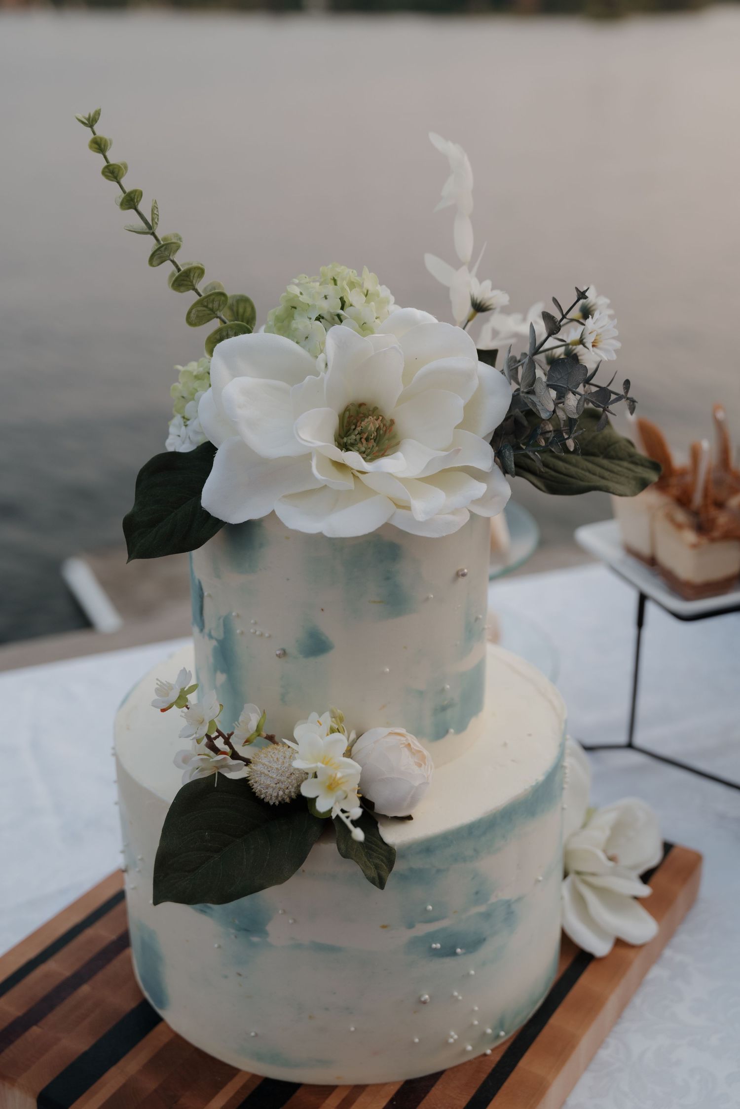 Elegant white wedding cake decorated with fresh white magnolias and eucalyptus on a wooden board.