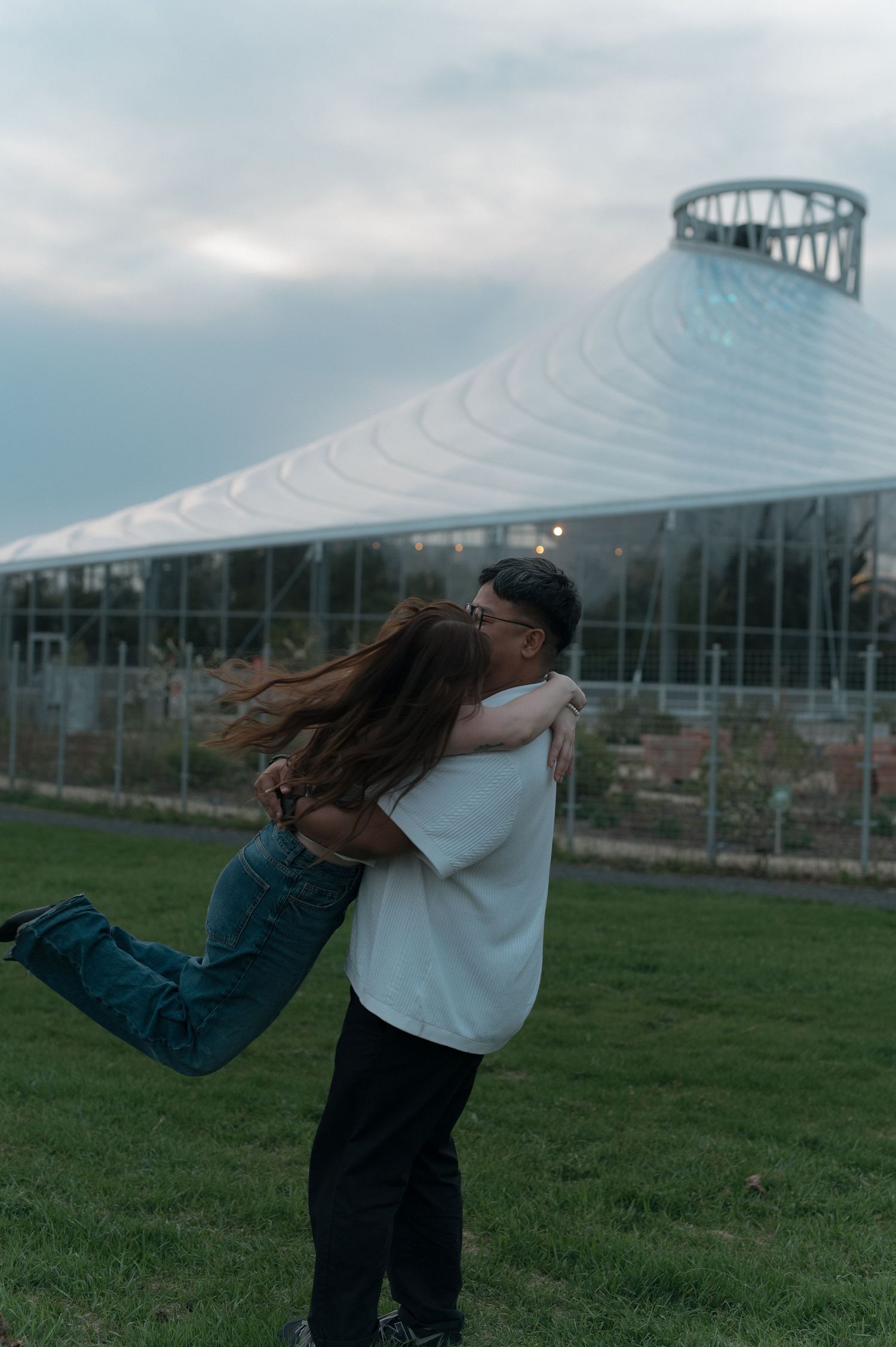 Couple sharing a romantic lift and kiss outdoors near a modern greenhouse structure under cloudy skies.