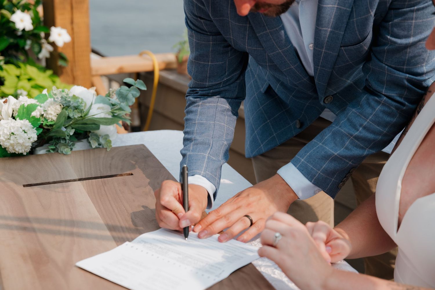 Hands signing wedding documents on a wooden table during an outdoor ceremony.
