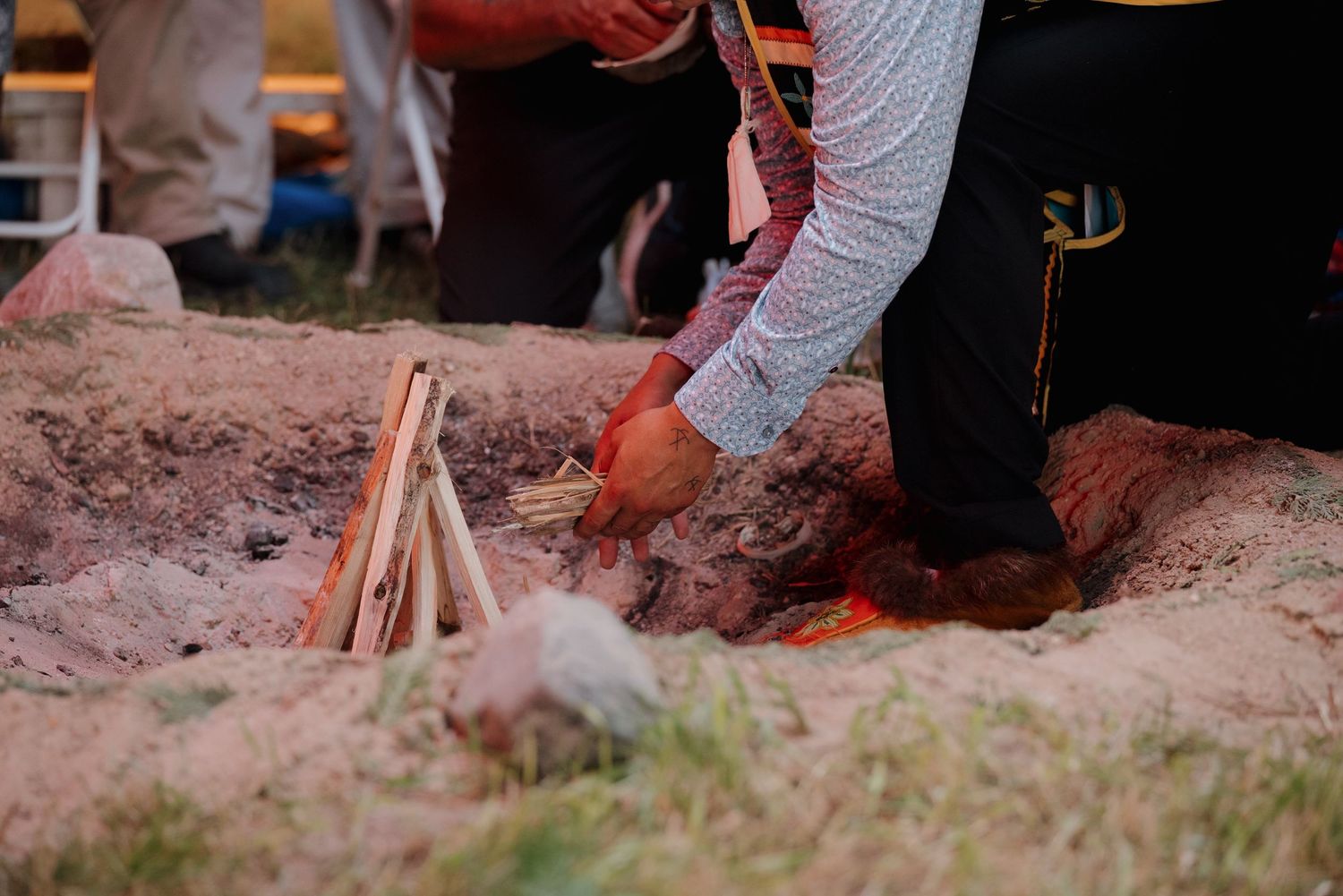 People digging in sandy soil at an outdoor cultural ceremony, wearing traditional orange and black attire at dusk.