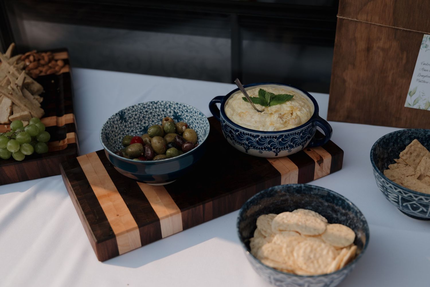 A rustic wooden serving board displays Mediterranean appetizers including hummus and olives in blue ceramic bowls.