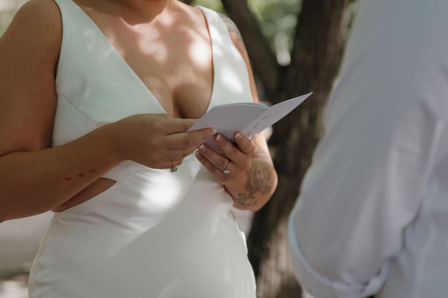 Bride in white dress reading wedding vows from a card during outdoor ceremony.