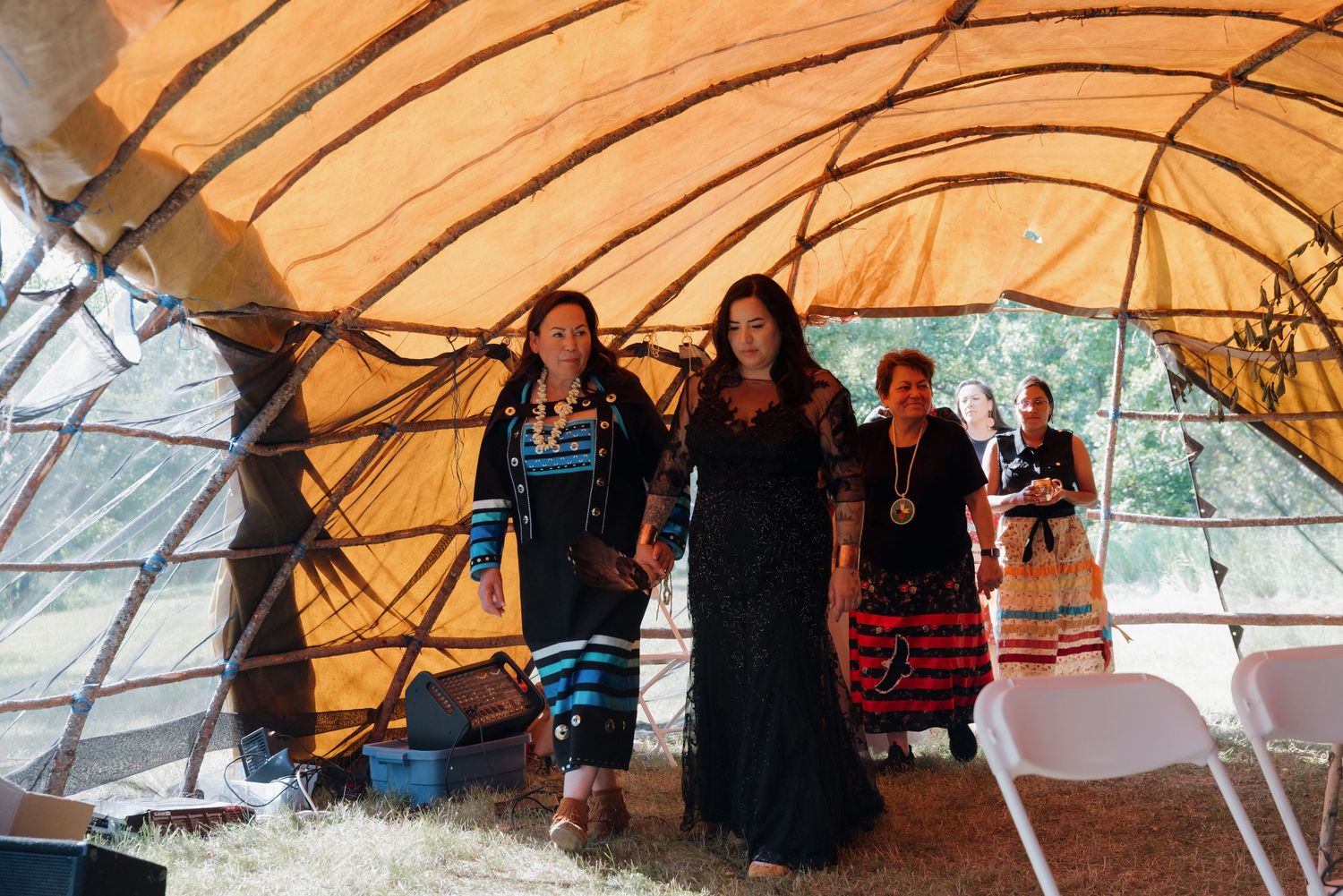 Three women in traditional Native American regalia stand together under a large ceremonial tent during a cultural gathering.